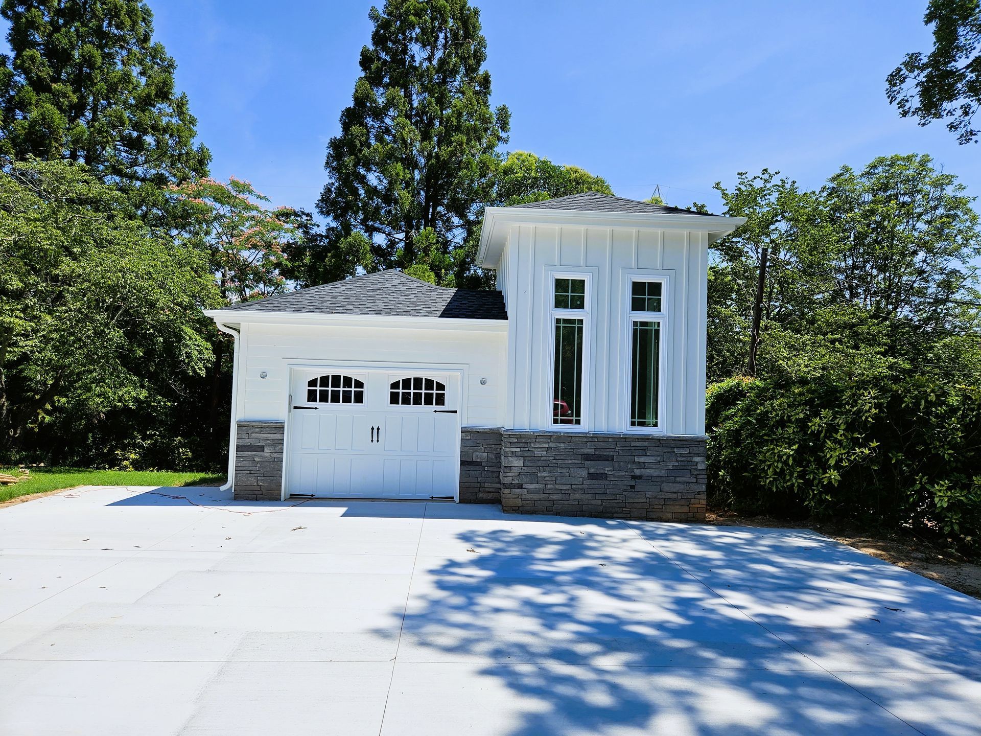 White garage with gray stone accents and tall windows, set amidst green trees under a blue sky.