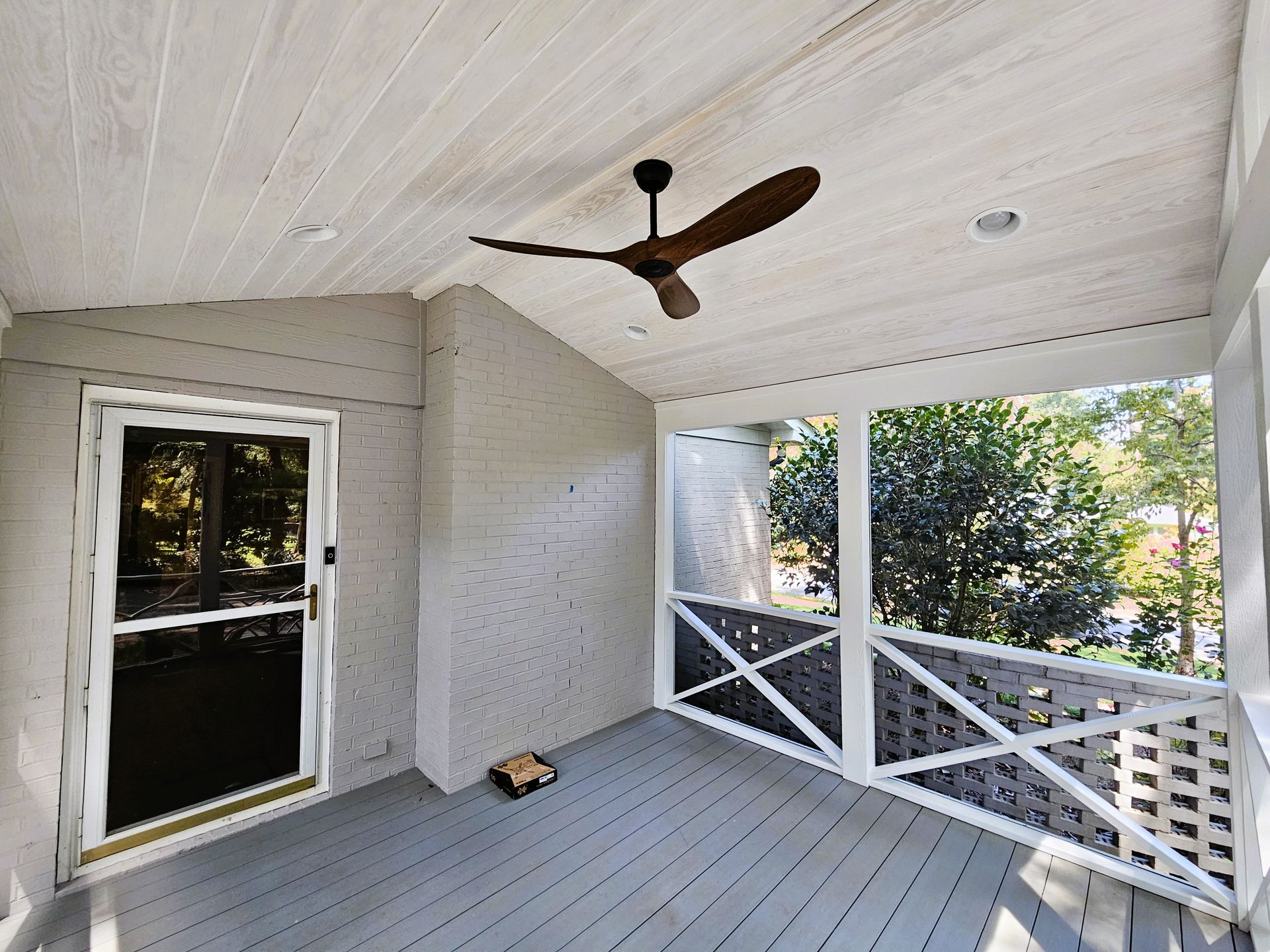 Covered porch with ceiling fan, door, and railing overlooking trees. Gray and white colors.
