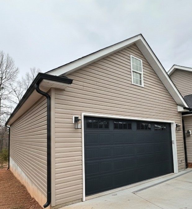 Tan-sided garage with black garage door, gutters, and trim, small window, cloudy sky.