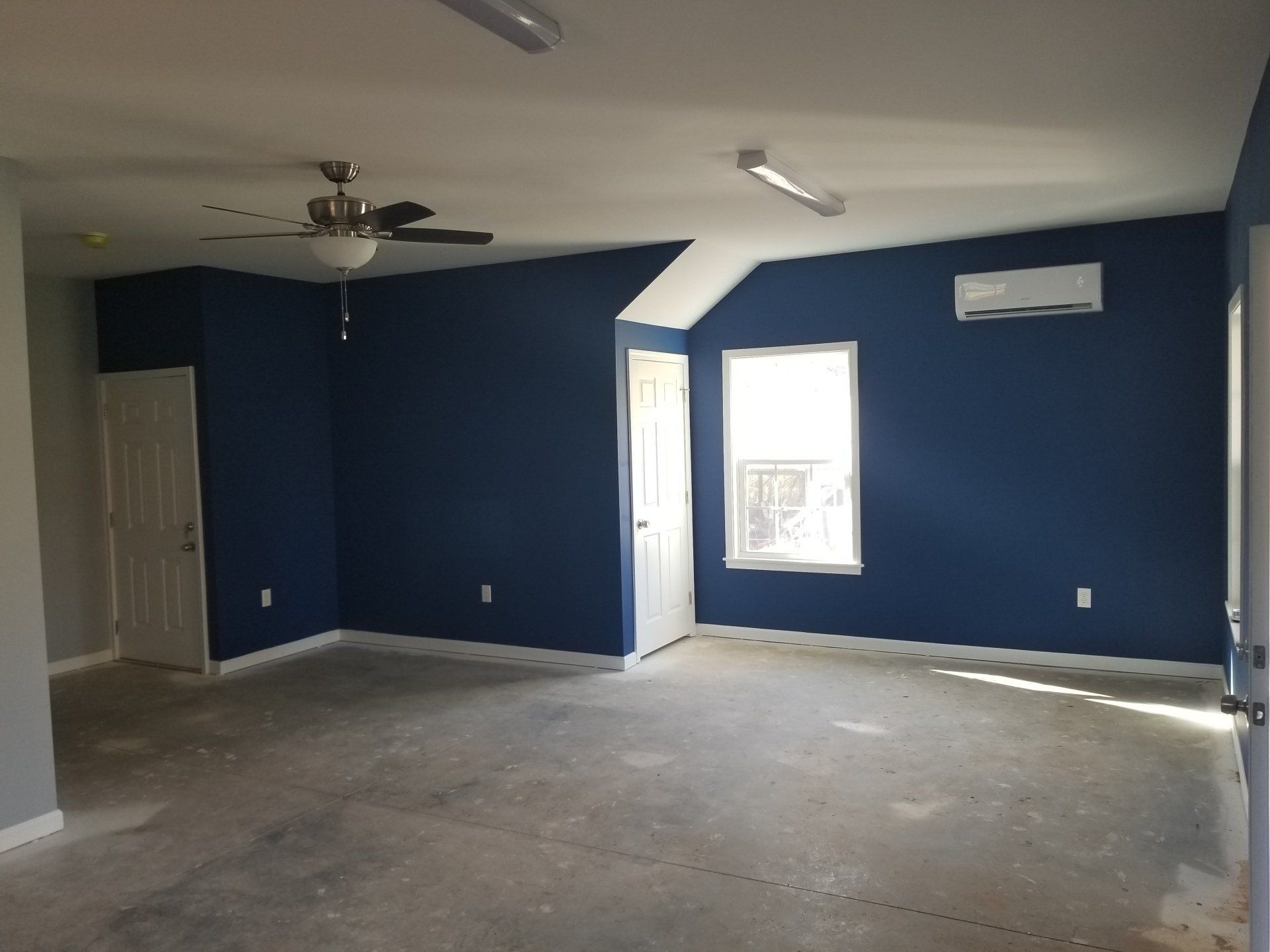 Empty room with blue walls, white ceiling, and a concrete floor. Includes a door, window, and ceiling fan.