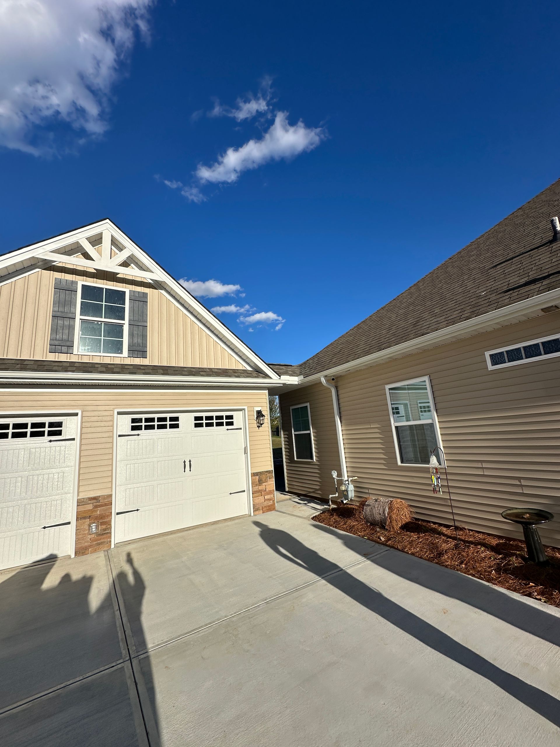 Exterior view of a beige house with a driveway. White garage doors, blue sky, and a shadow are visible.