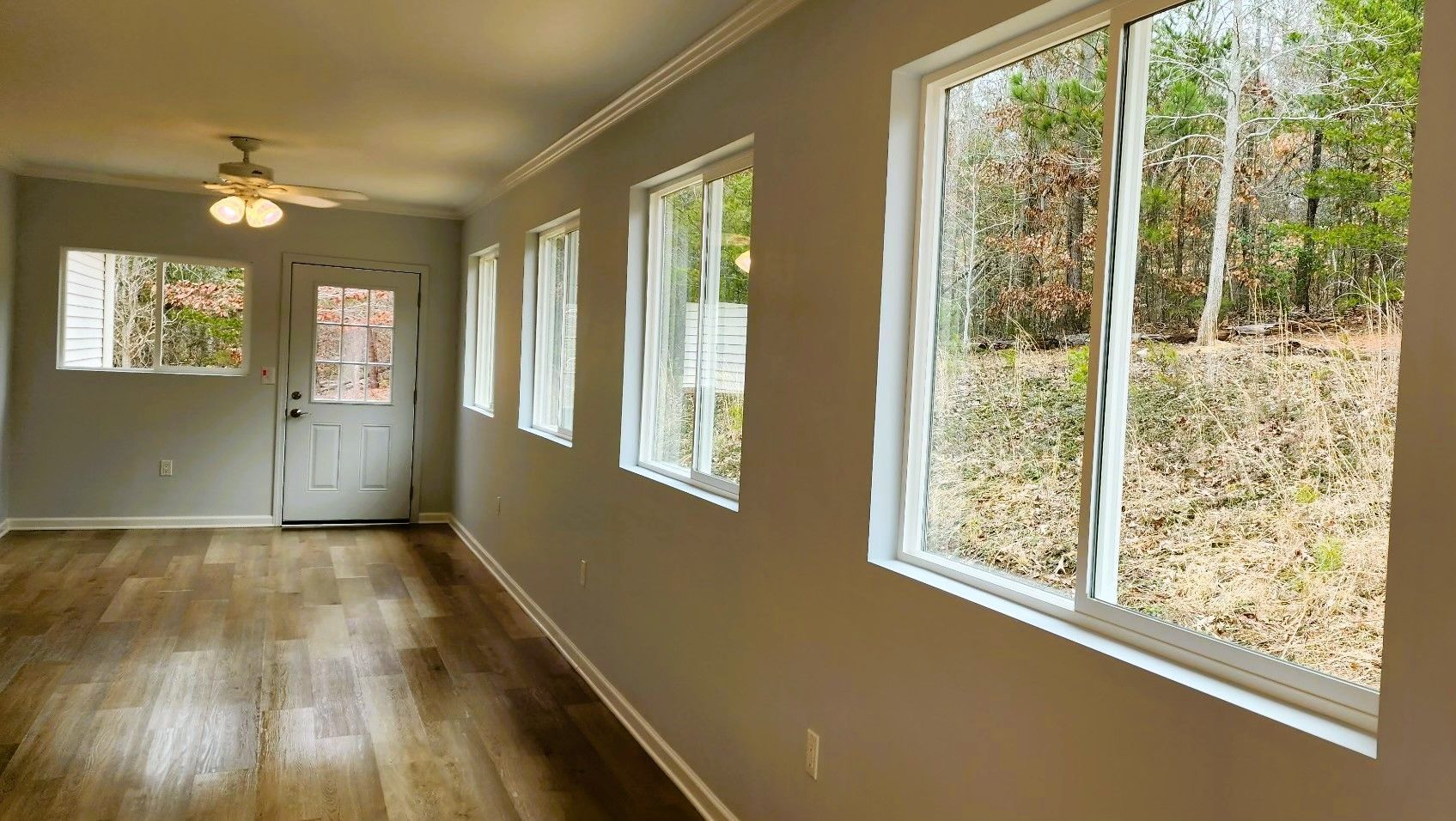 Interior view of a room with wood-look flooring, gray walls, and multiple windows overlooking a wooded area.