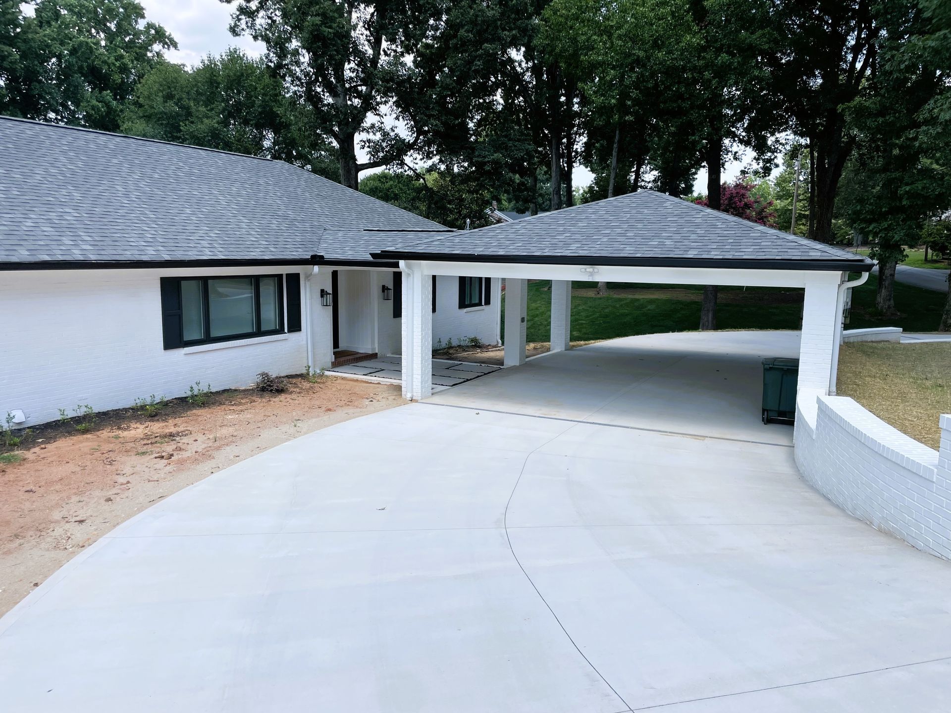 White house with a carport; gray roof, white walls, concrete driveway, and trees.