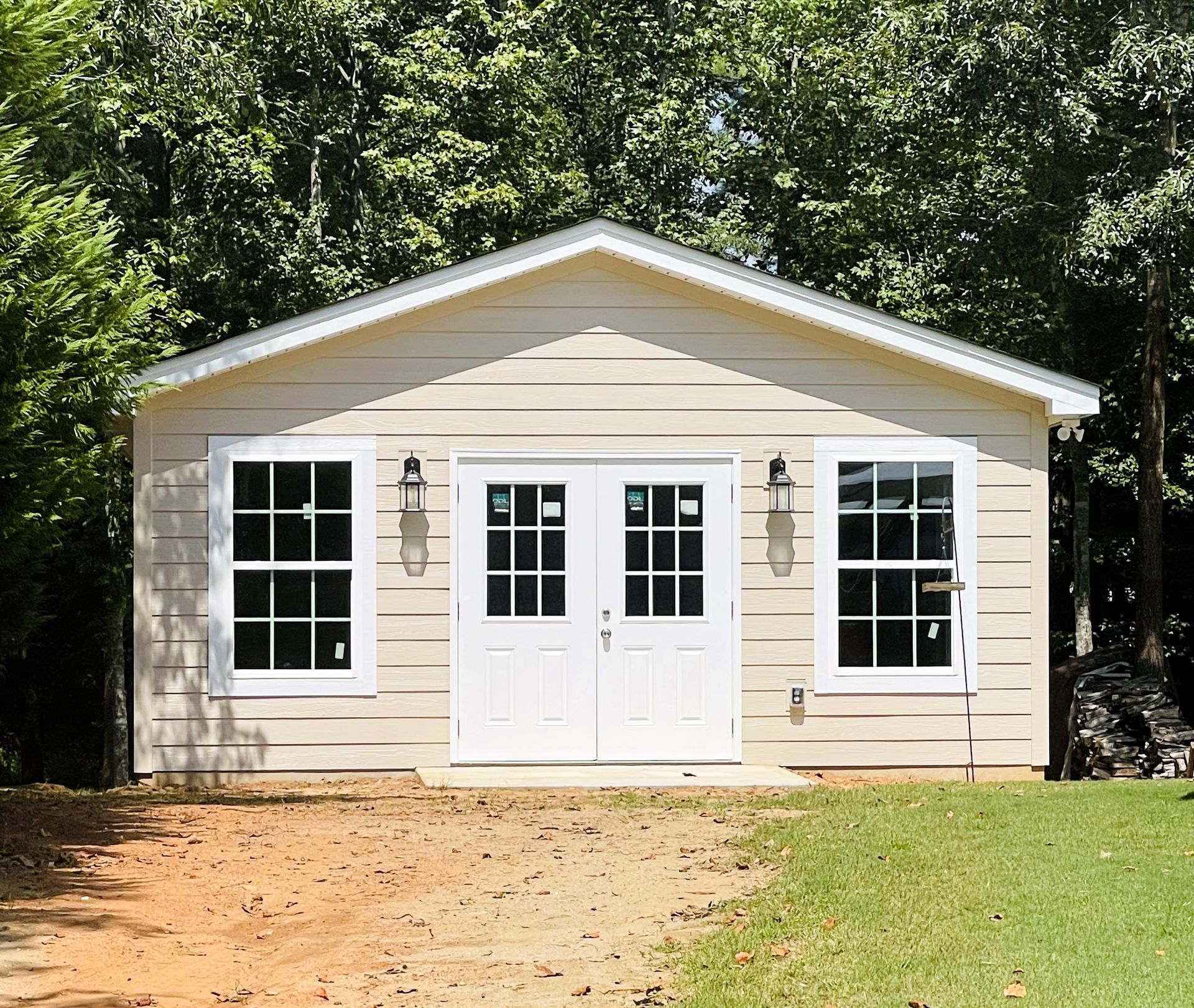 Beige shed with white double doors and windows, set against a backdrop of trees.