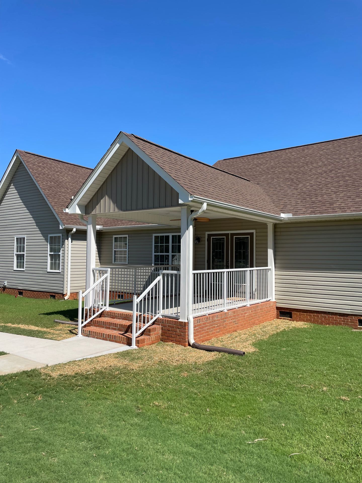 A light gray house with a brown roof and a porch with white railings and brick accents under a blue sky.