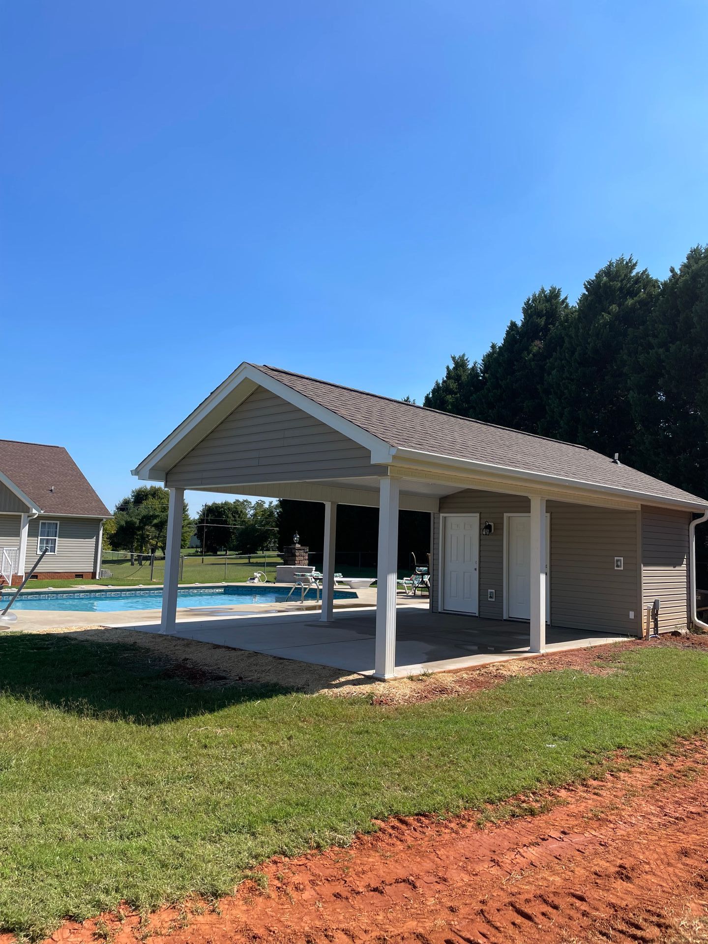 Poolside cabana with tan siding, white pillars, and a brown roof. Includes a pool, building, and trees.