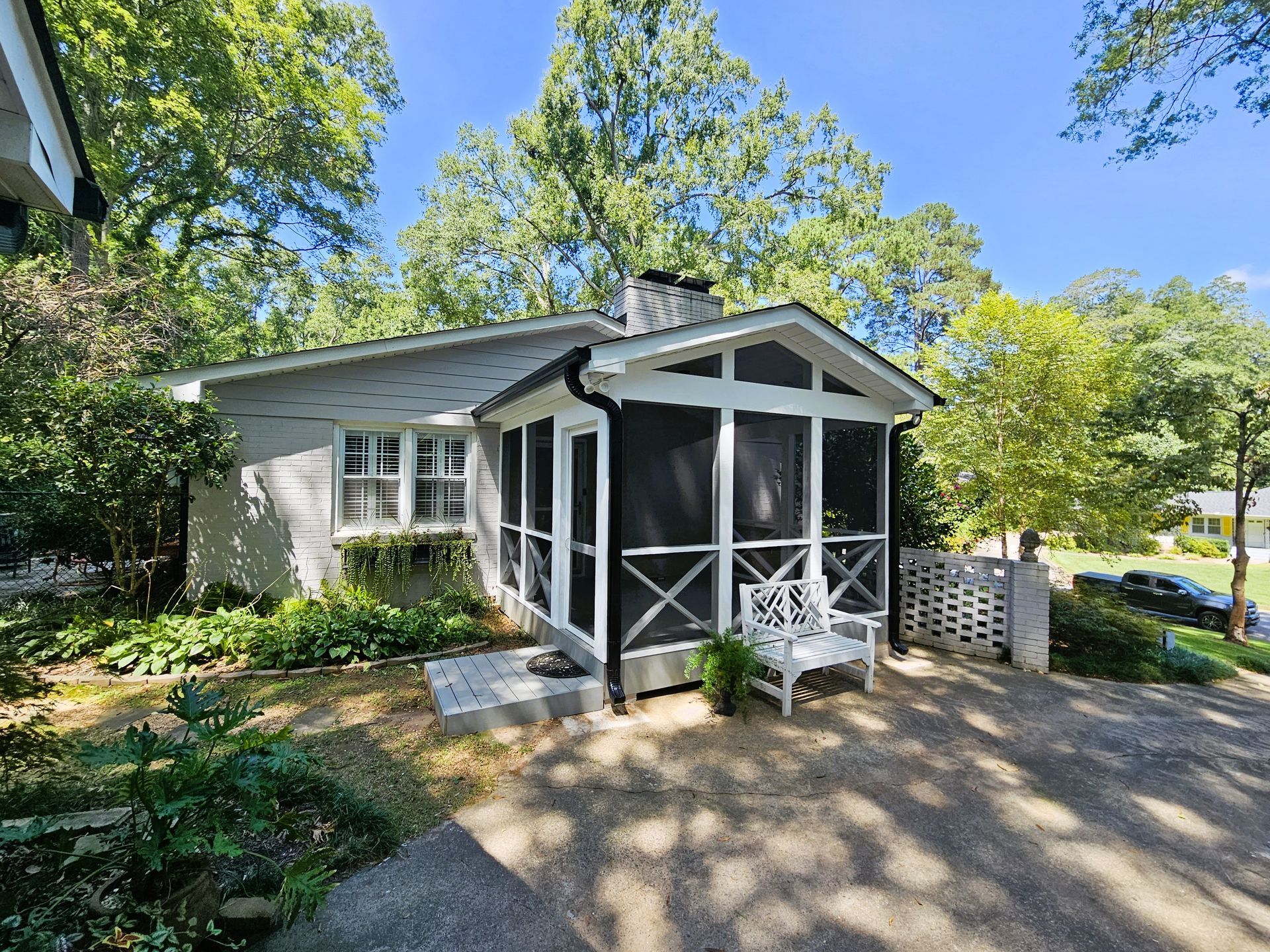 Small gray cottage with screened porch, white trim, and a bench outside. Surrounded by greenery.