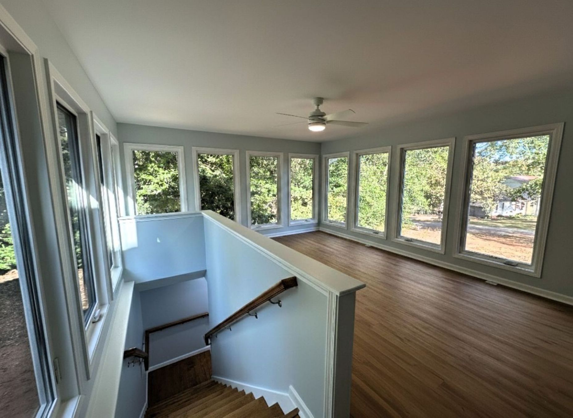 Sunroom with numerous windows, wooden floors, and a staircase. Light blue walls, white trim, and a ceiling fan.