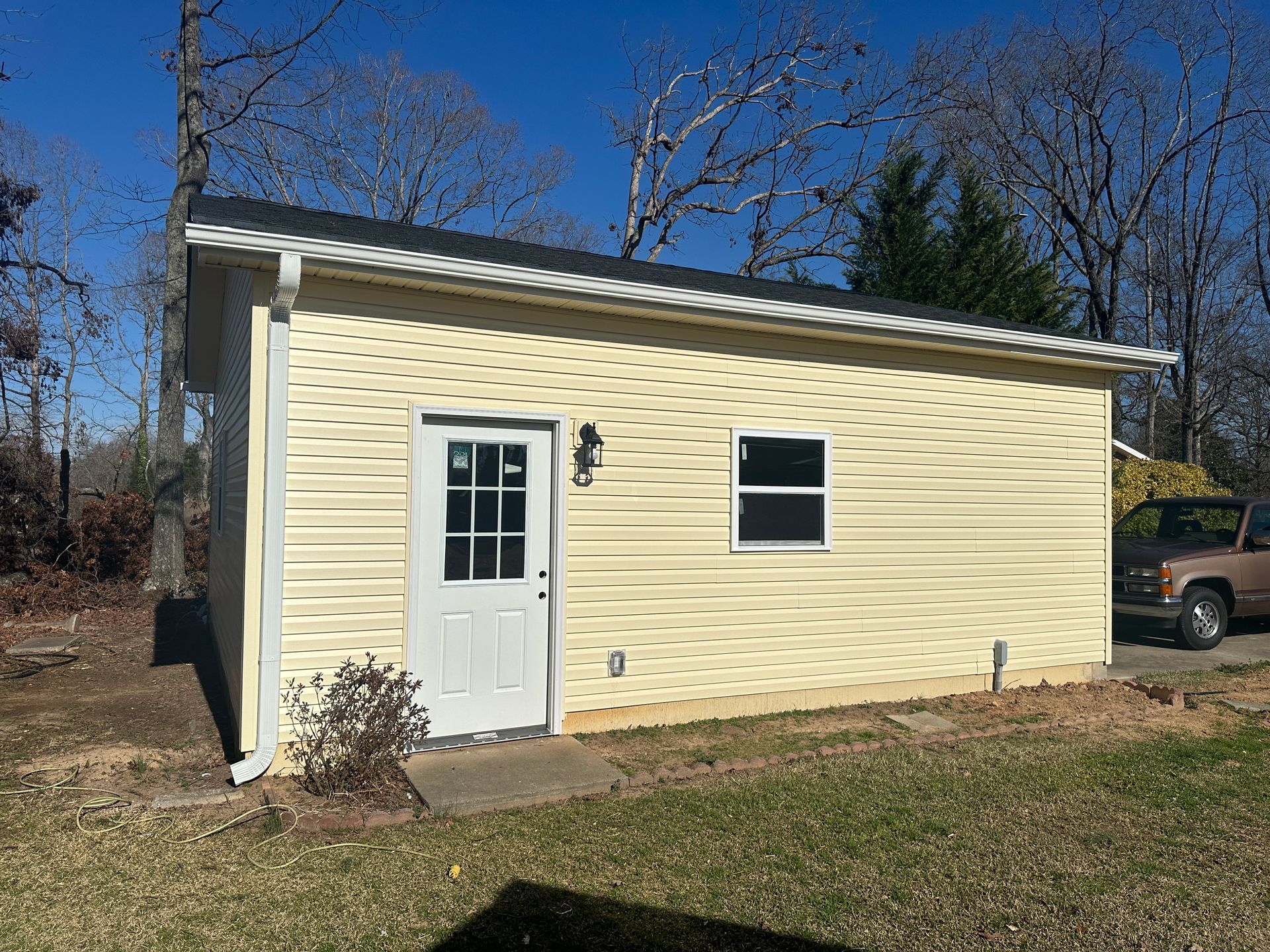 Yellow-sided shed with white door and window, dark roof, situated in a grassy yard, with a clear blue sky.