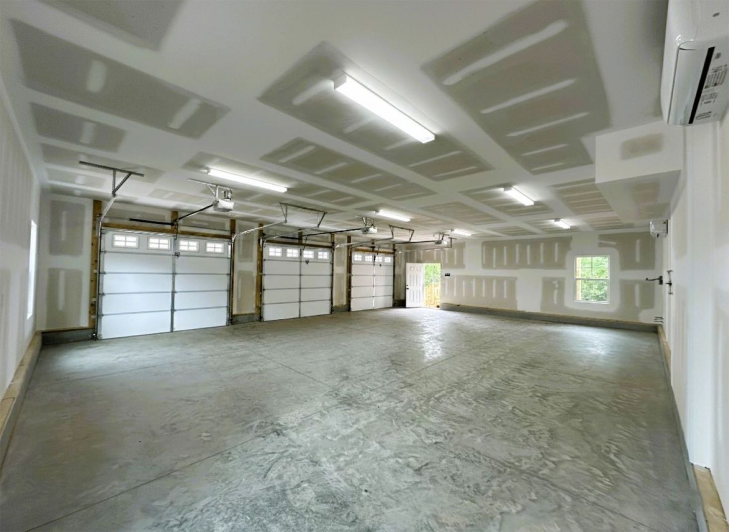 Empty, unfinished garage interior with three garage doors and exposed drywall. Fluorescent lights are on.