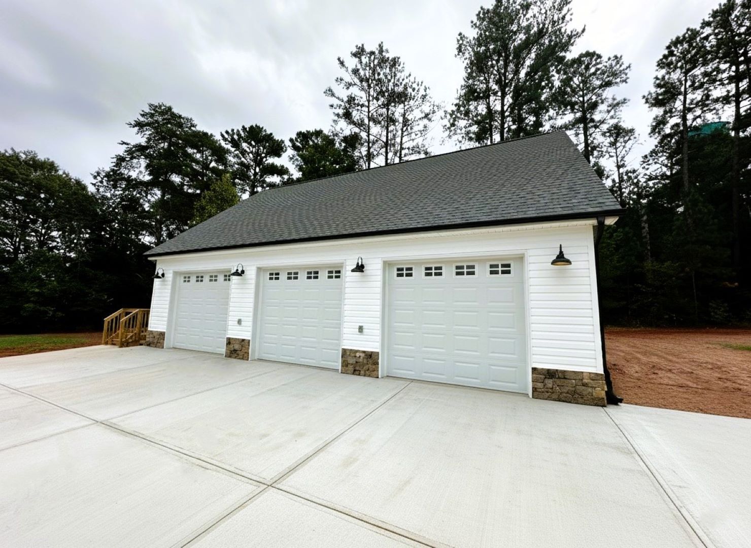 Three-car garage with white siding, gray doors, and a concrete driveway. Trees are in the background.
