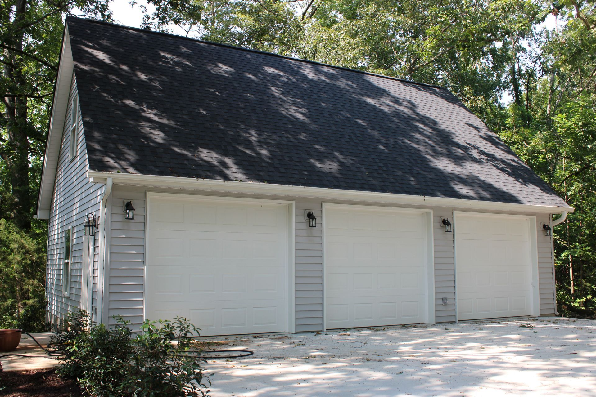 Three-car garage with white doors and gray siding, black roof, set against trees.