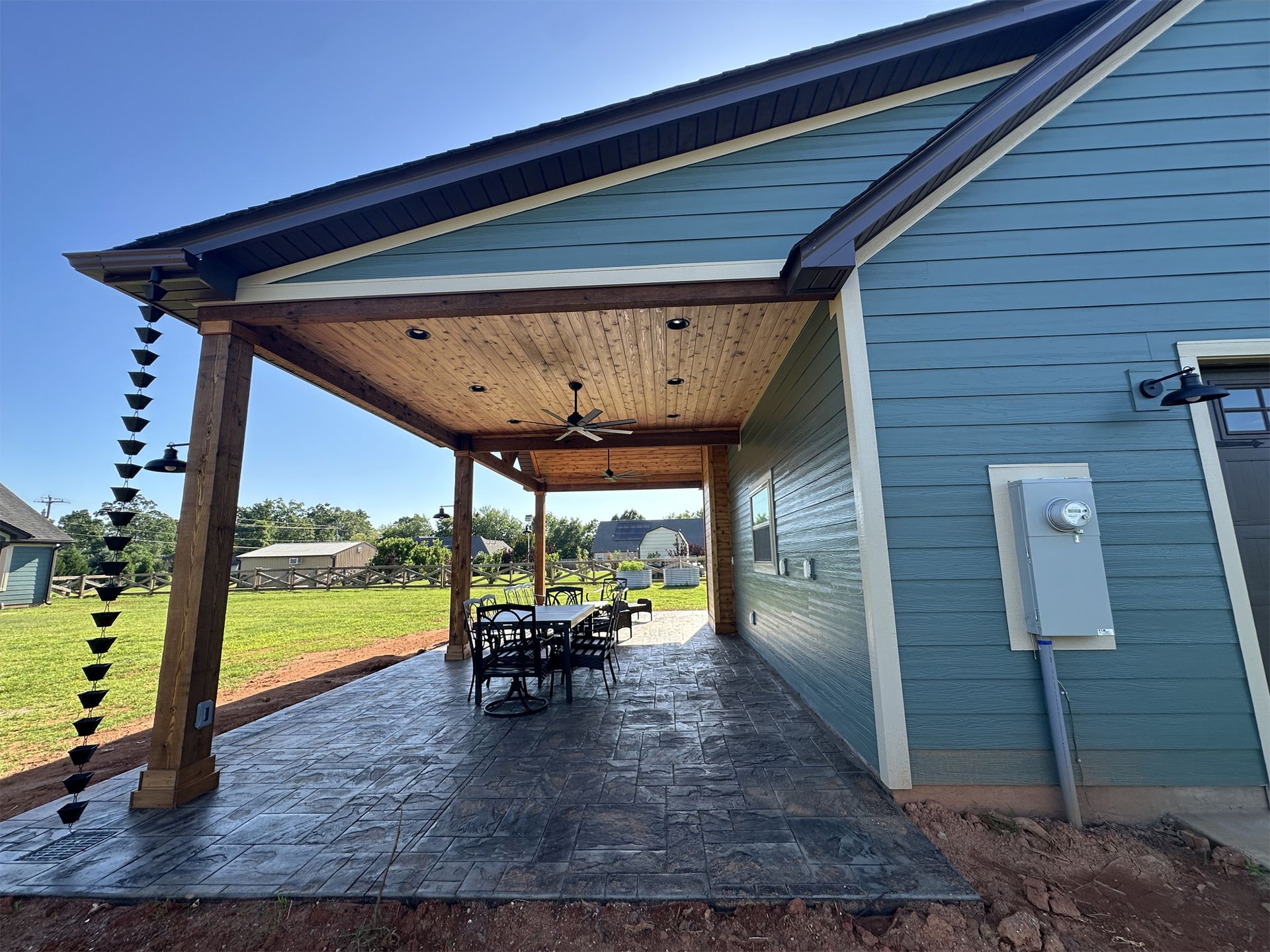 Covered patio with wooden supports, gray stamped concrete floor, blue siding, rain chain.