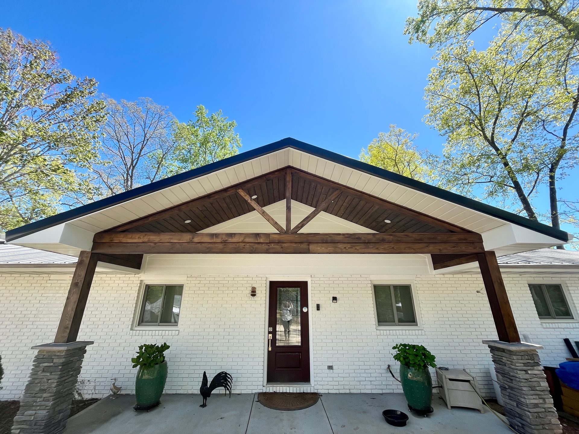 White house with brown wooden porch, stone pillars, and green plants under a clear blue sky.