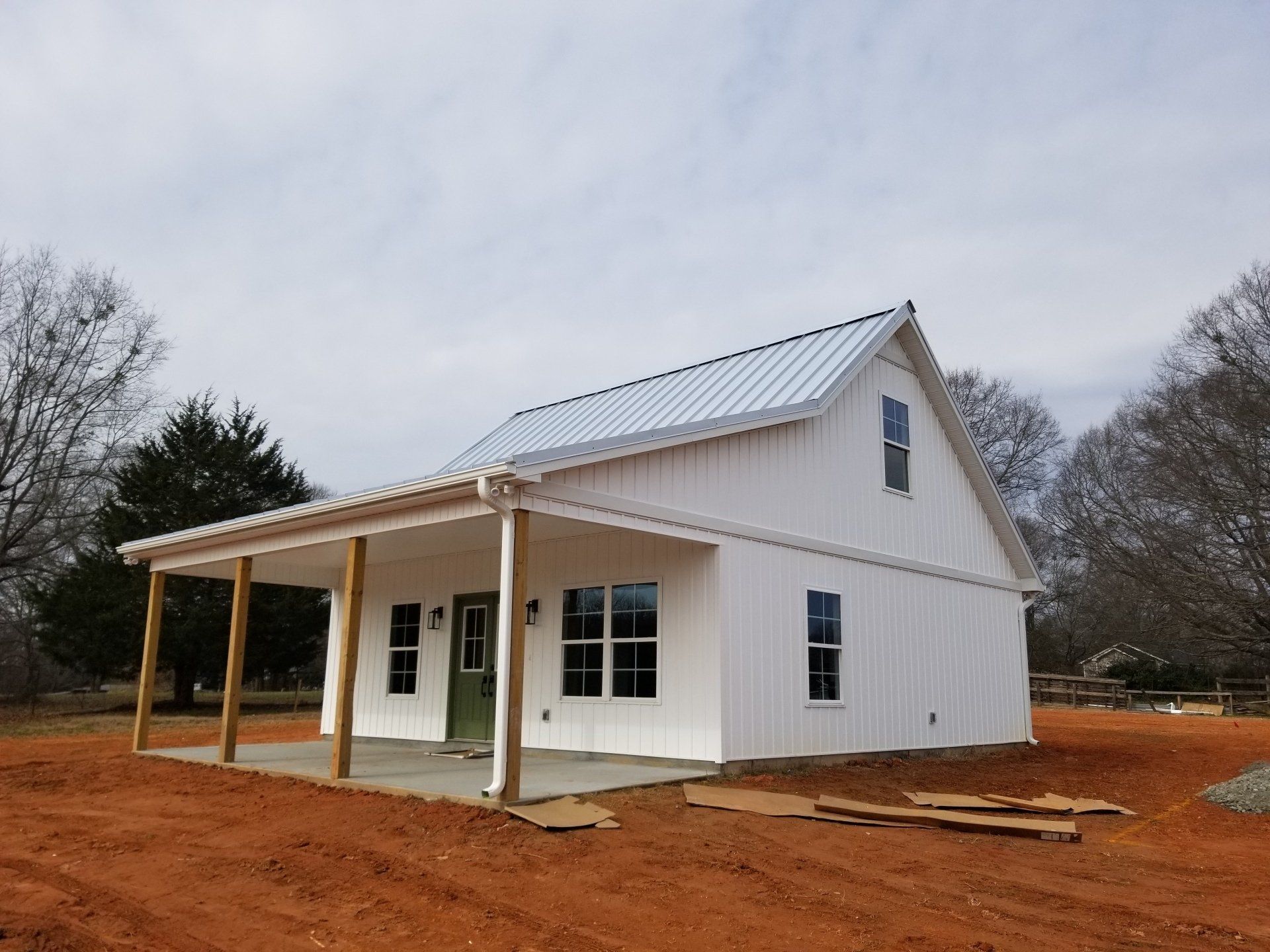 White two-story building with a porch, metal roof, and green door on a red dirt lot under a cloudy sky.