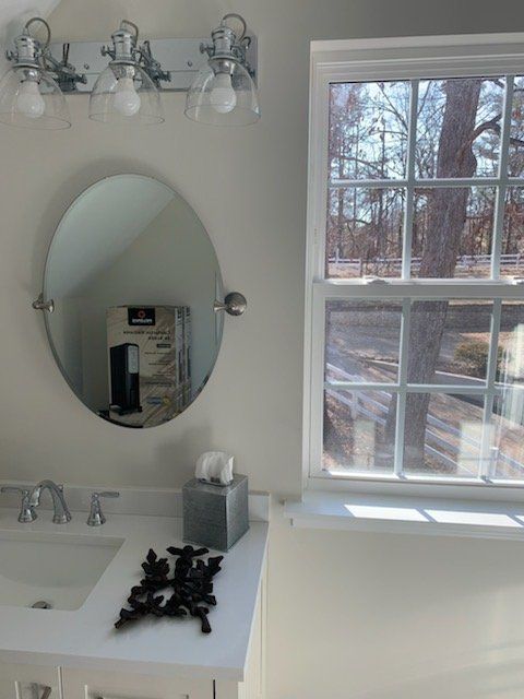 Bathroom with oval mirror, vanity, and window overlooking trees. White walls, chrome fixtures.
