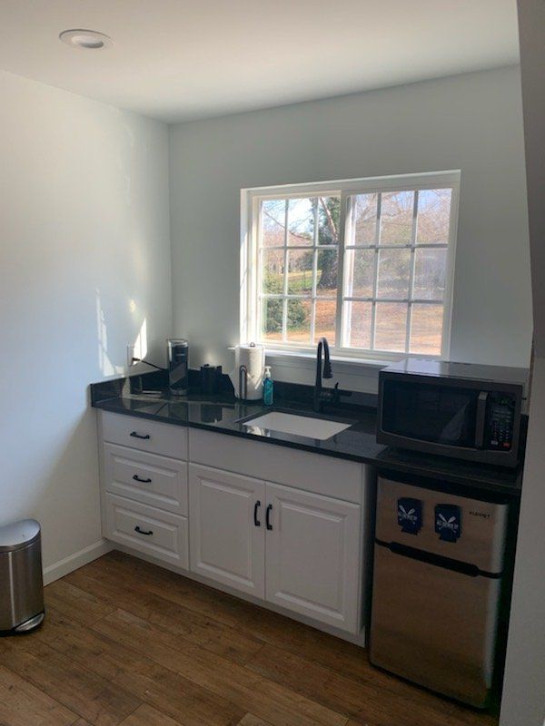 Small kitchen with white cabinets, black countertop, sink, window, and mini-fridge.