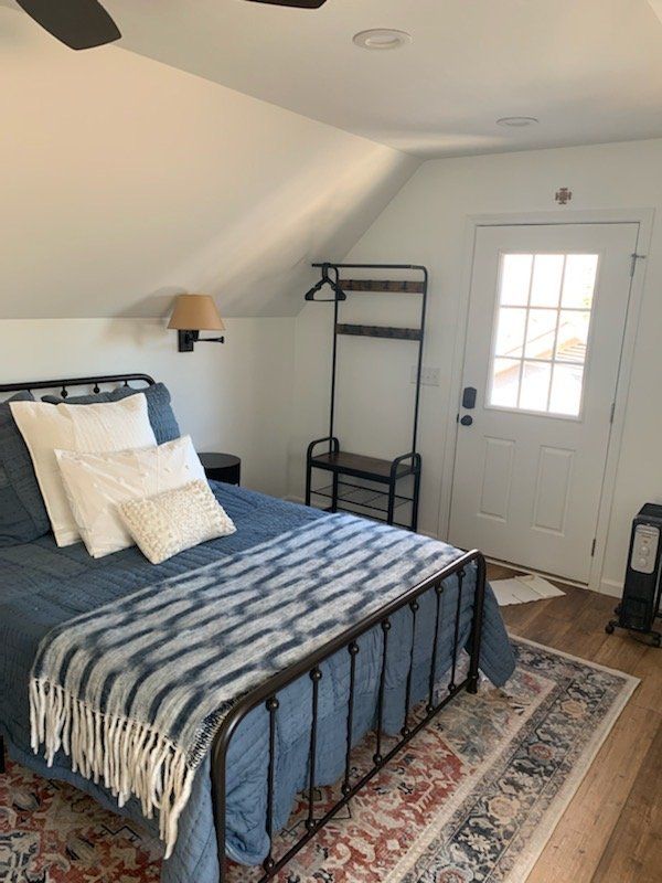 Bedroom with a bed, rug, coat rack, and a door, in a room with white walls and wood flooring.