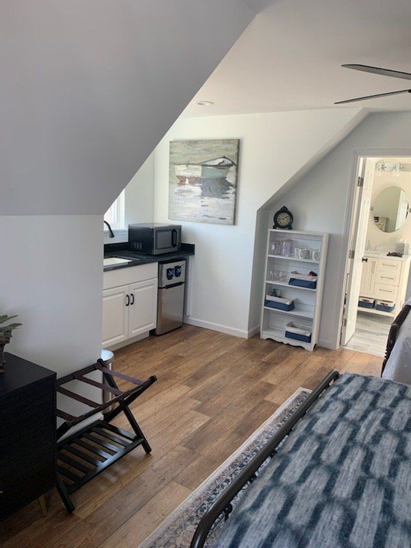 A small kitchenette area with a microwave, mini-fridge, and sink. White cabinets, wood-look floor, and art. Doorway to a bathroom.