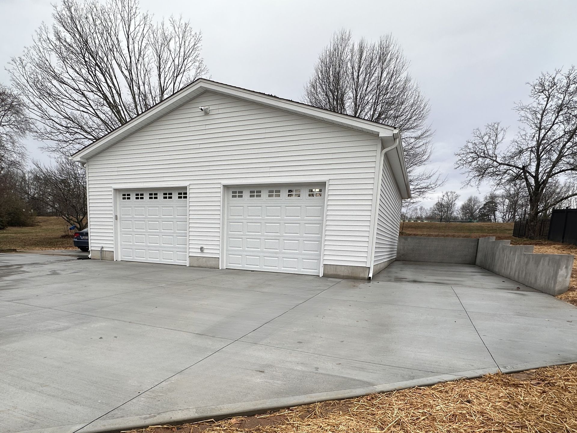 A white two-car detached garage with a concrete driveway and a low concrete retaining wall in a rural, overcast setting.