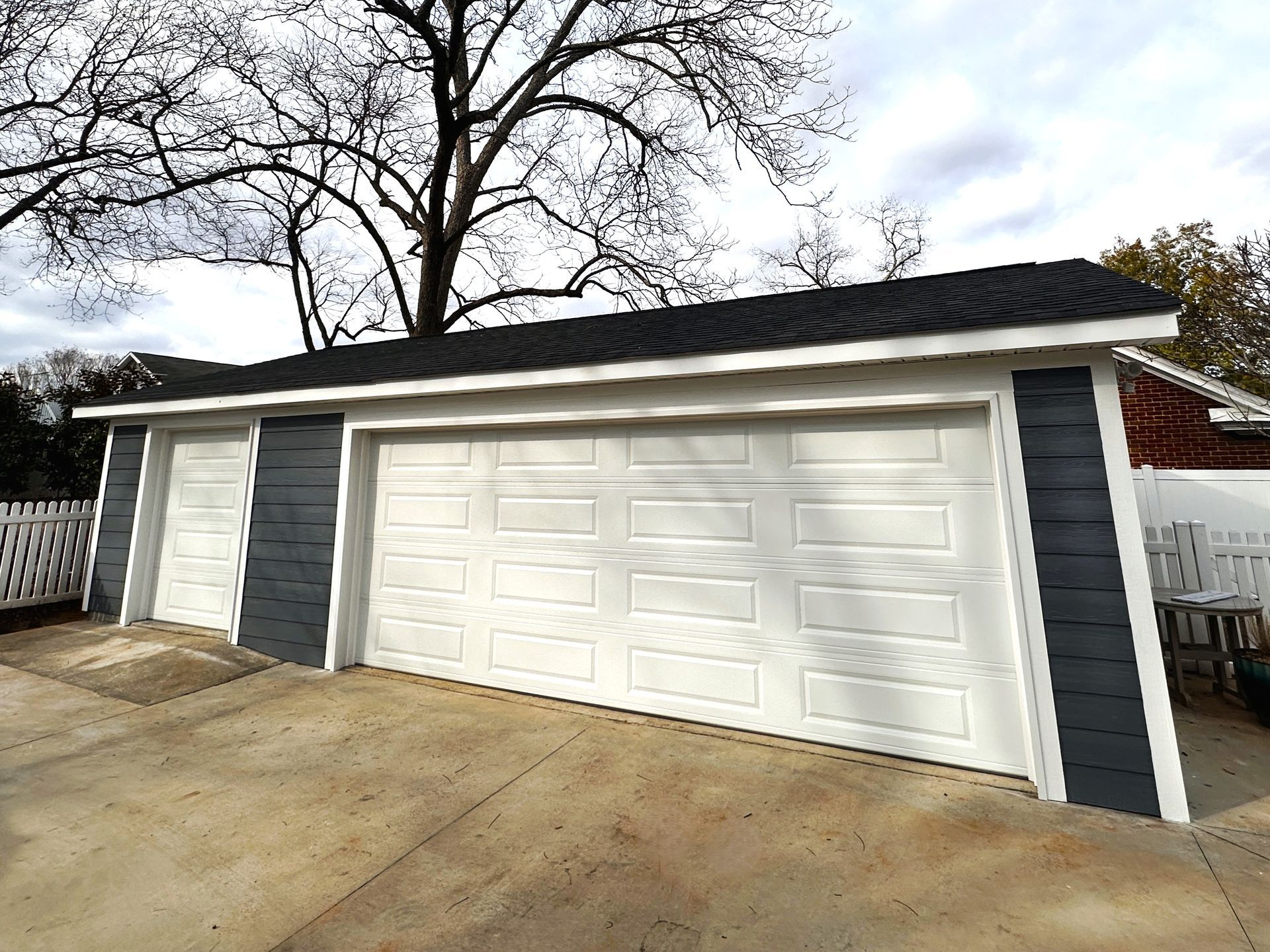 A detached garage featuring dark grey siding, white doors, and a black shingled roof under a clear, cloudy sky.