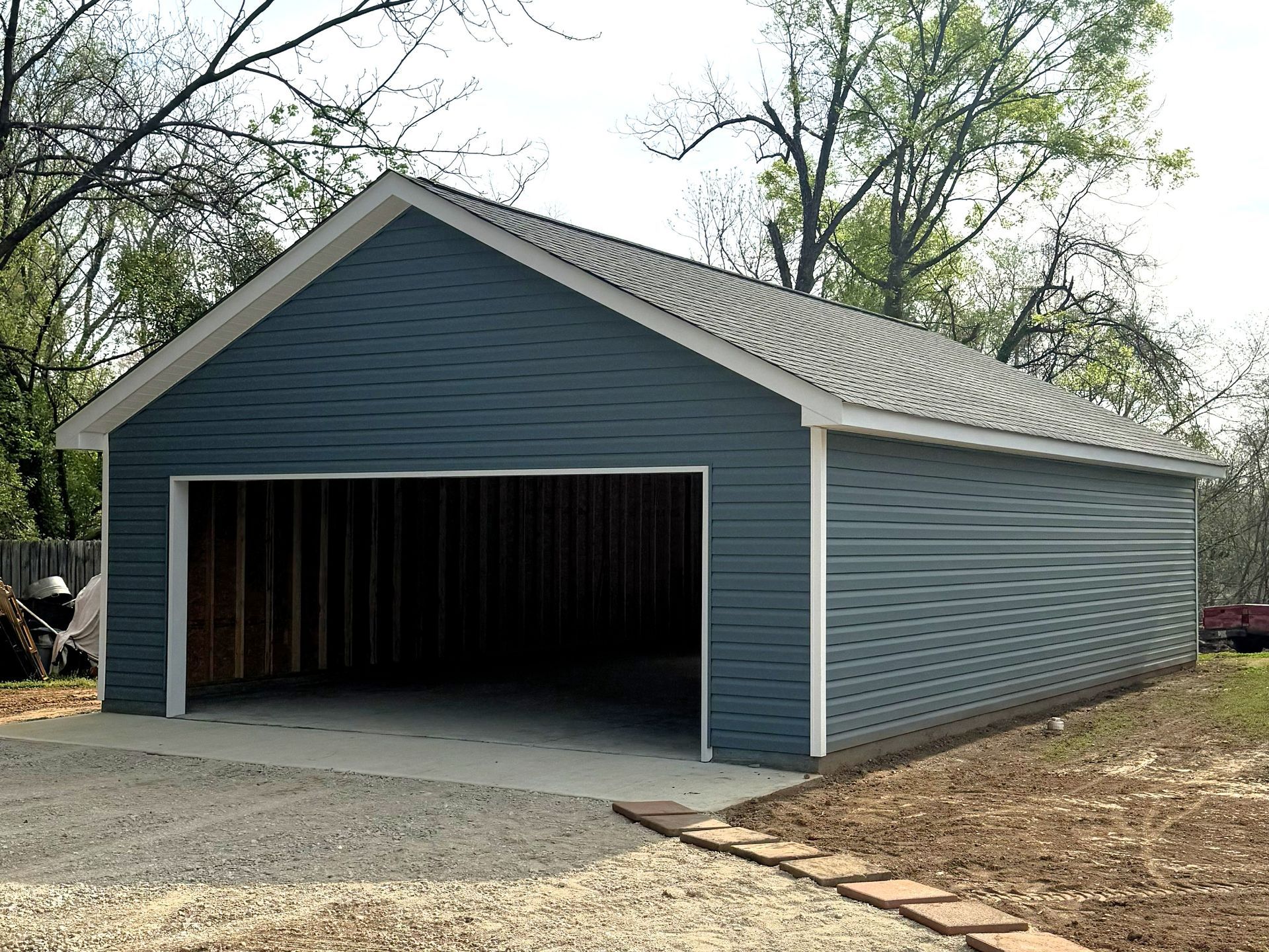 A blue-sided garage with a large open entrance, asphalt roof, and white trim, situated on a gravel lot with trees.