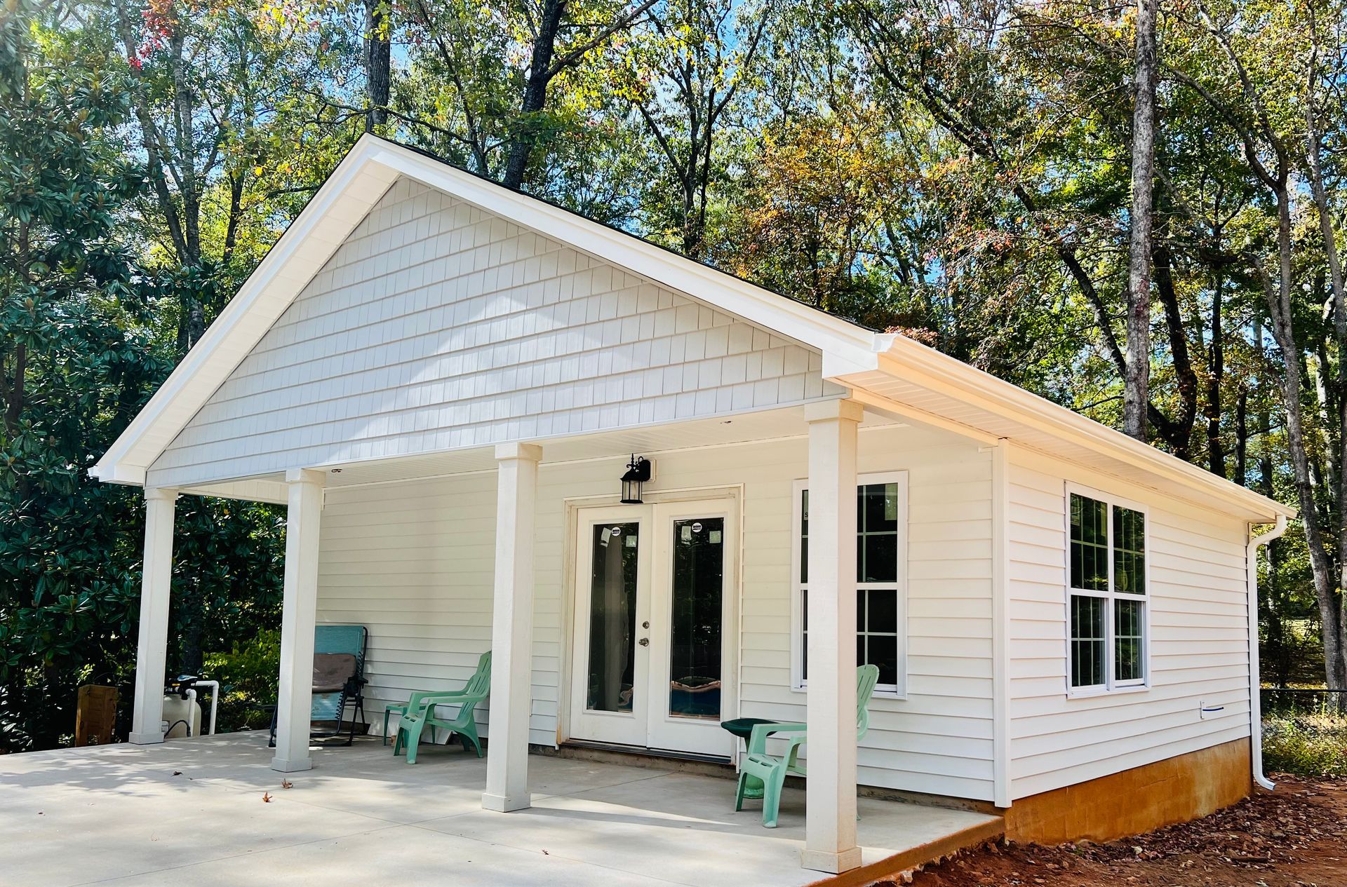 White cottage with porch, two green chairs, surrounded by trees.