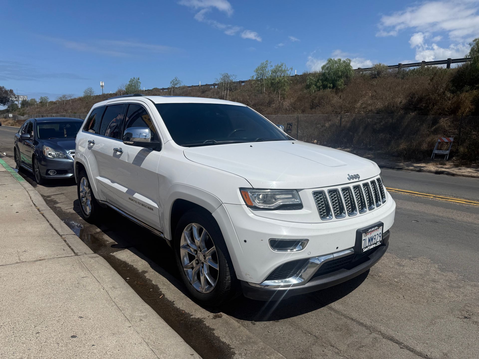 White Jeep SUV parked on a roadside with a black car behind it under a clear blue sky