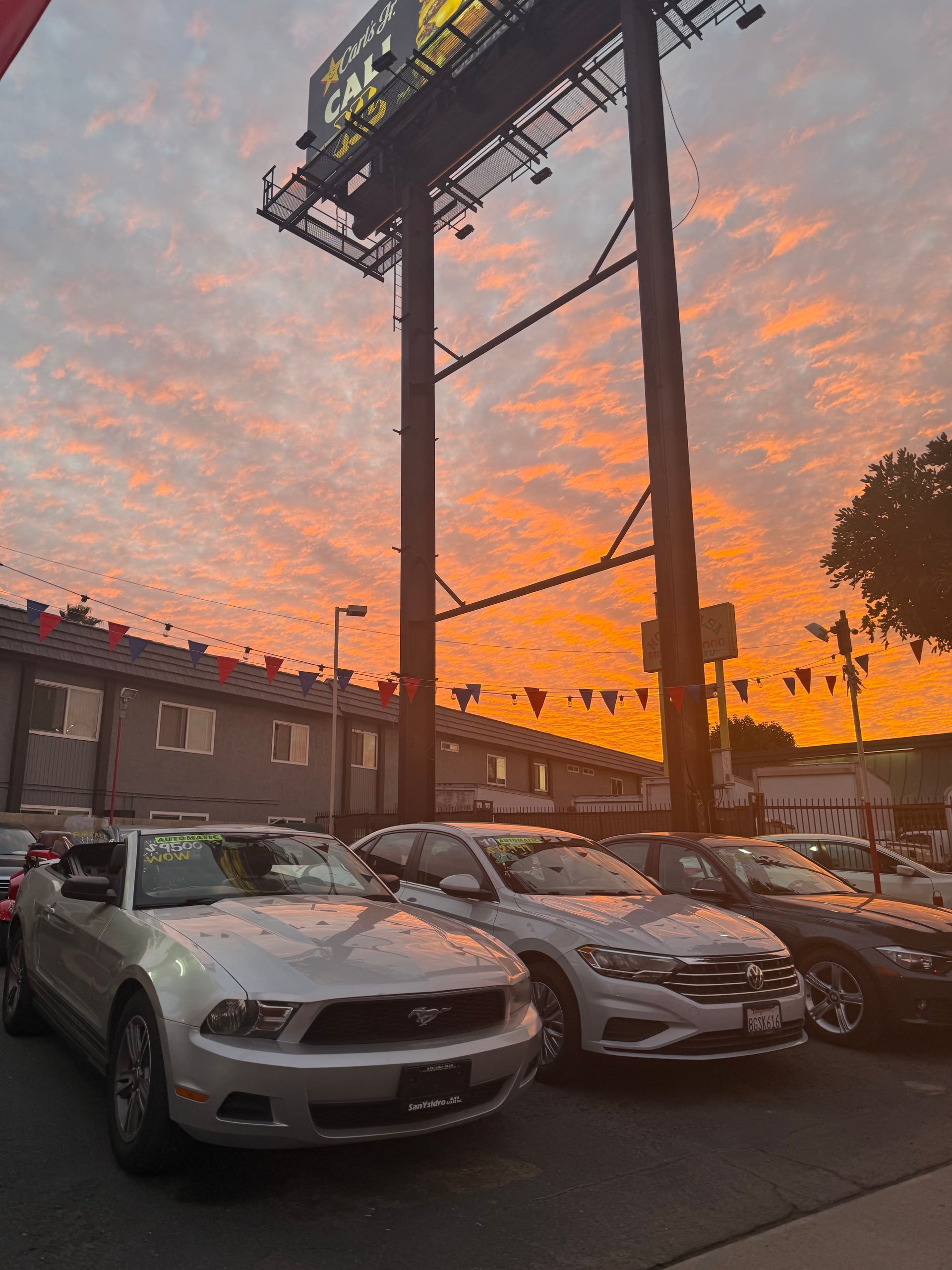 Cars parked under a billboard at sunset, with an orange sky and silhouetted utility poles.