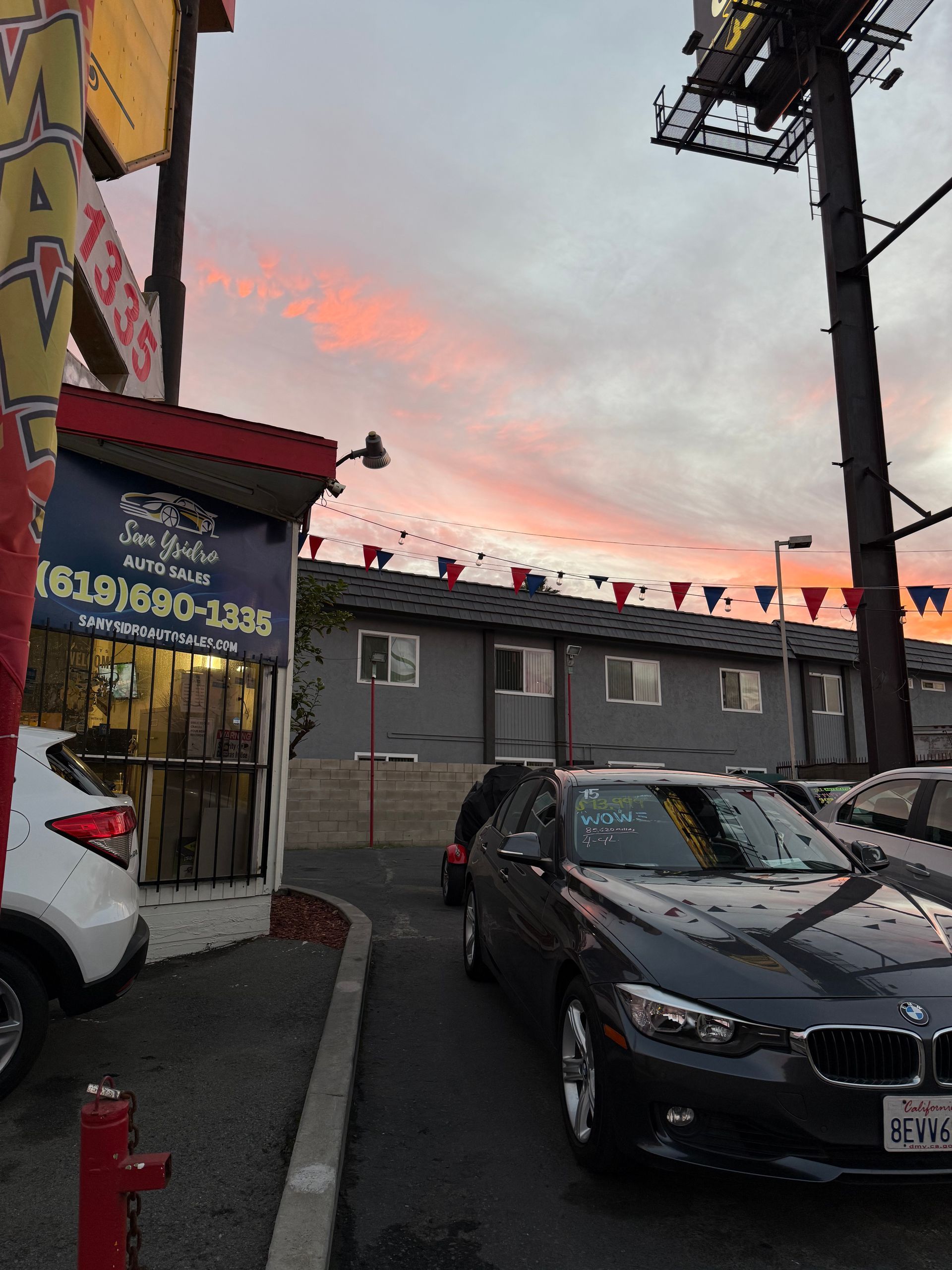Urban street at dusk with parked cars, colorful storefronts, and pink clouds in the sky.