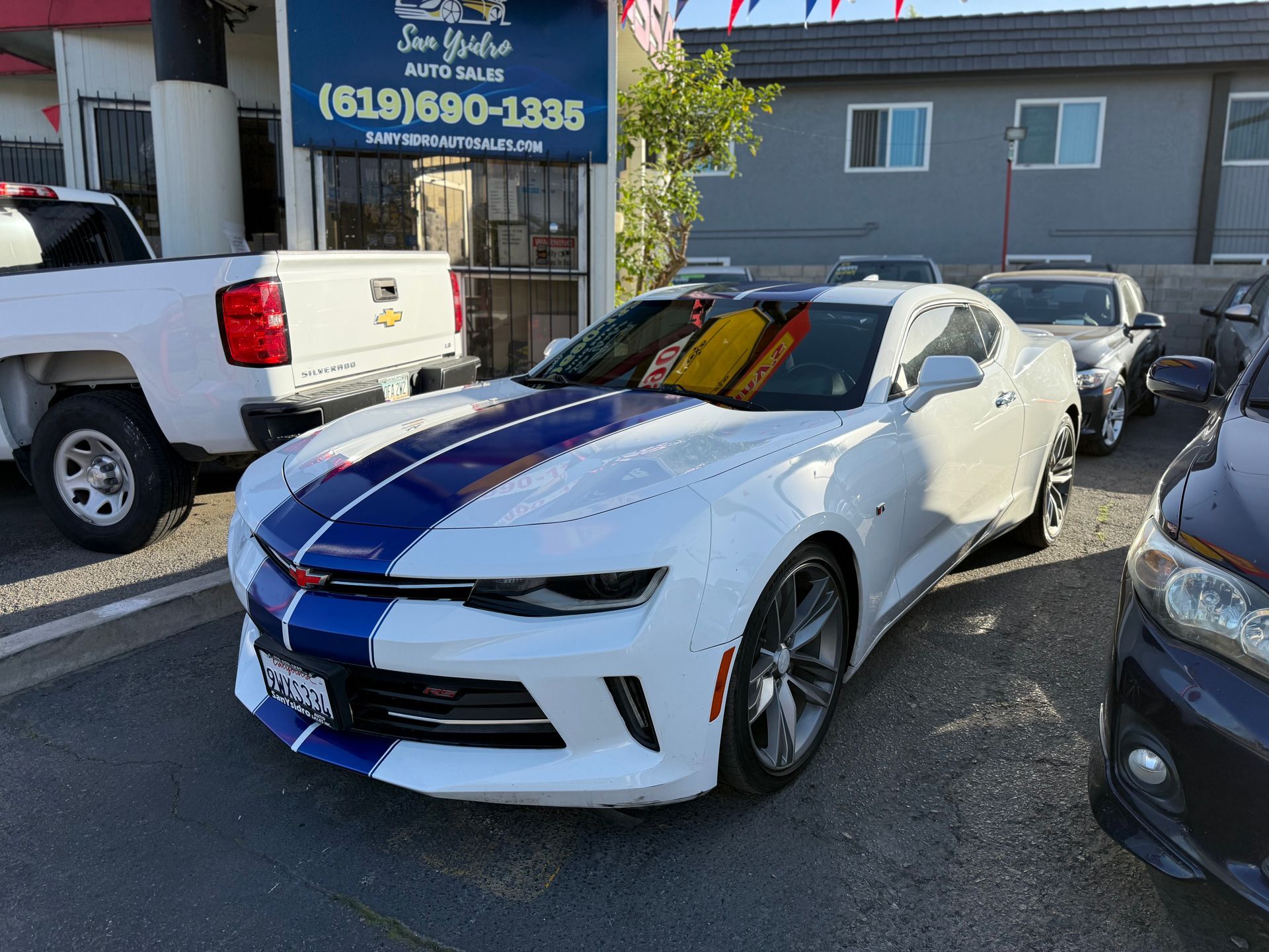 White sports car with blue racing stripes parked in a car lot beside other vehicles