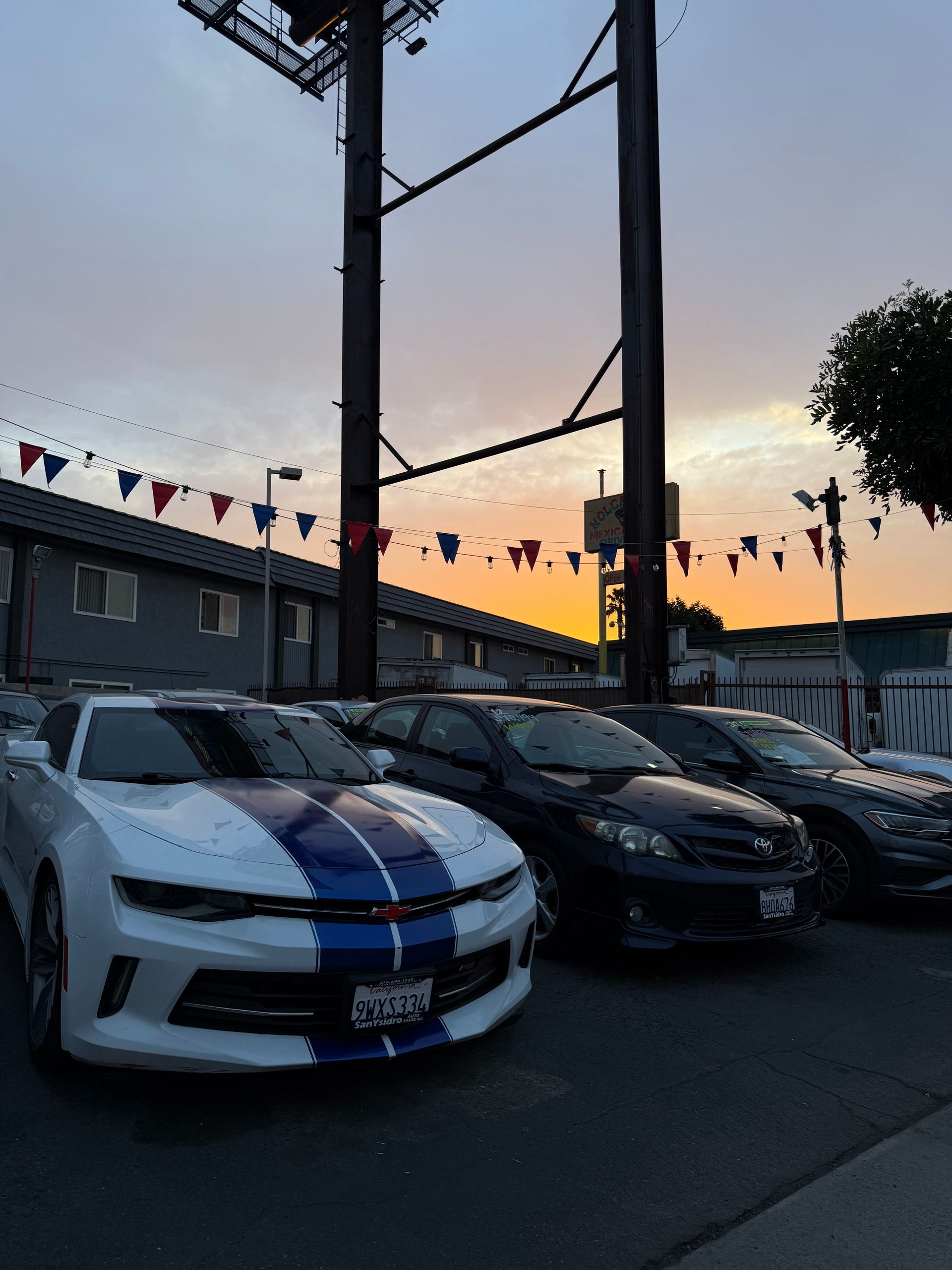 Parked cars, including a white sports car with blue stripes, lined up at dusk under pennant flags.