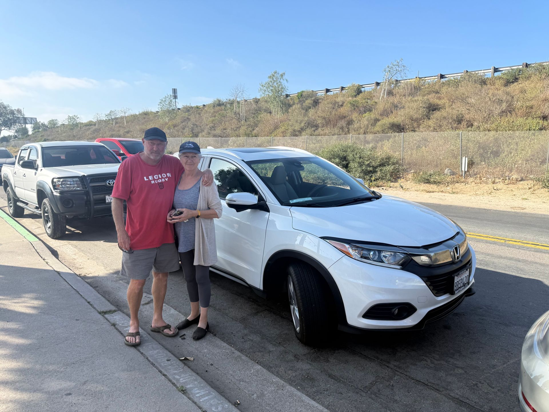 Two people standing beside a white SUV parked on a roadside with several cars behind them