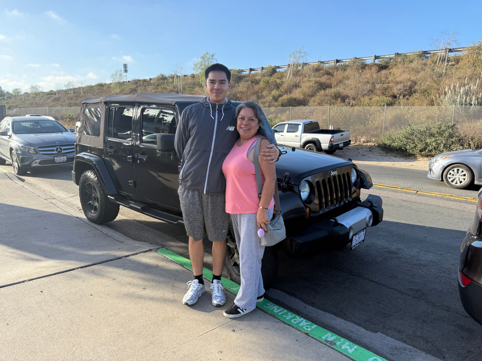 Two people standing in front of a black Jeep on a sunny roadside with hills in the background