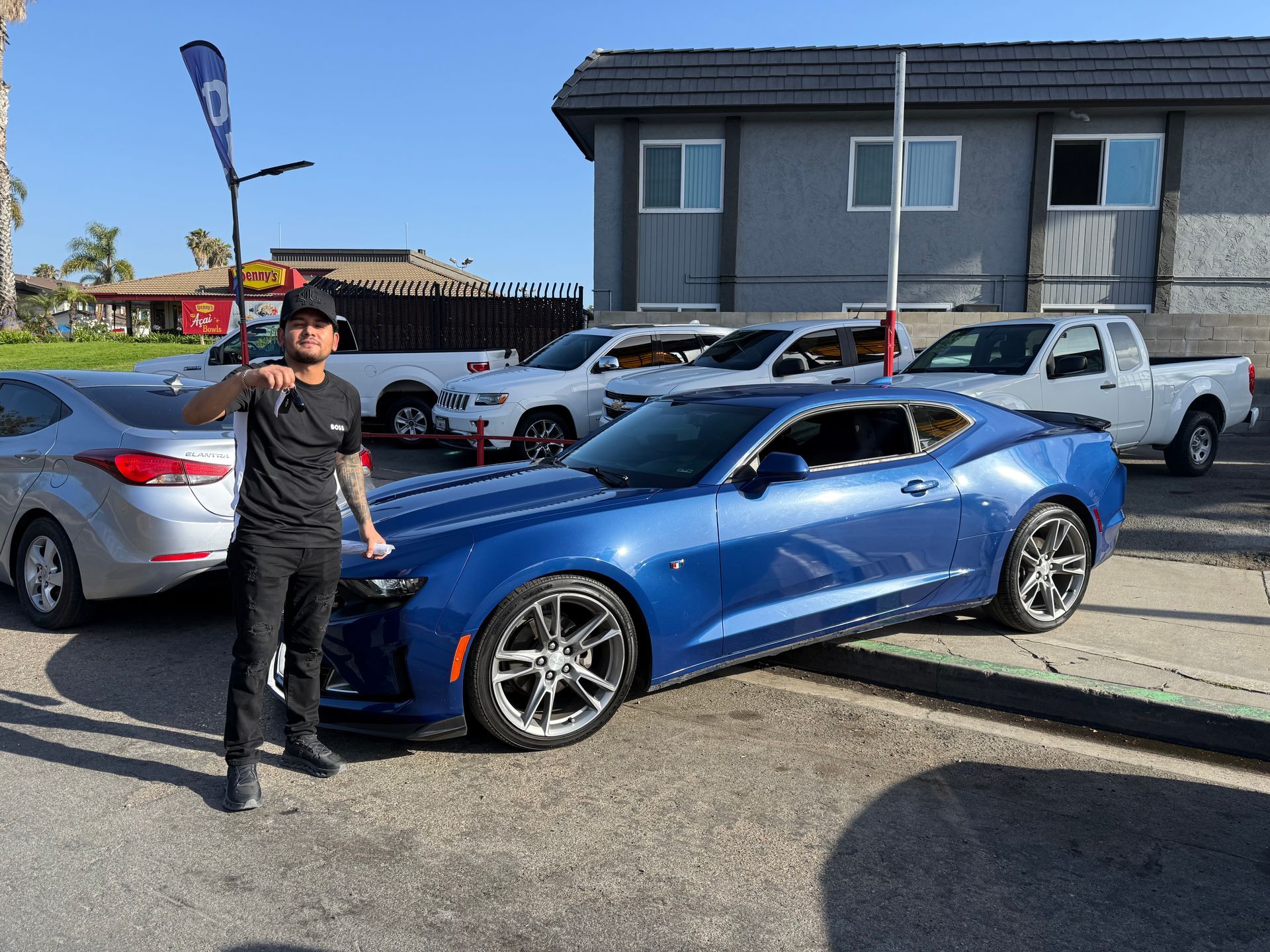 Person standing beside a blue sports car in a parking lot near a gray building.