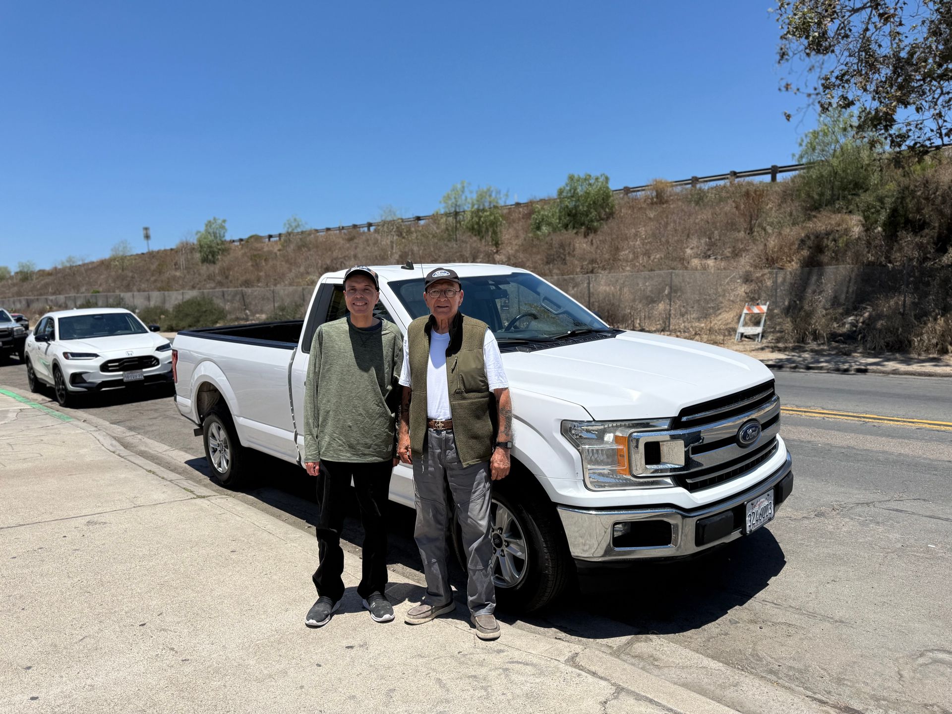 Two people standing beside a white pickup truck on a sunny roadside
