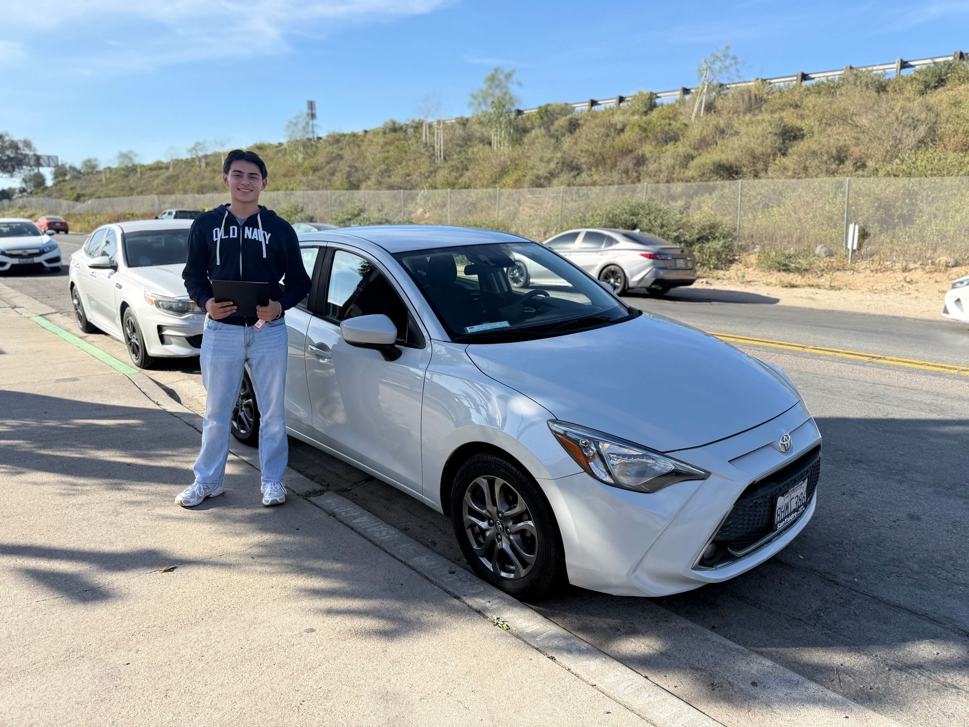 Person standing beside a white sedan on a roadside with parked cars and a hillside in the background