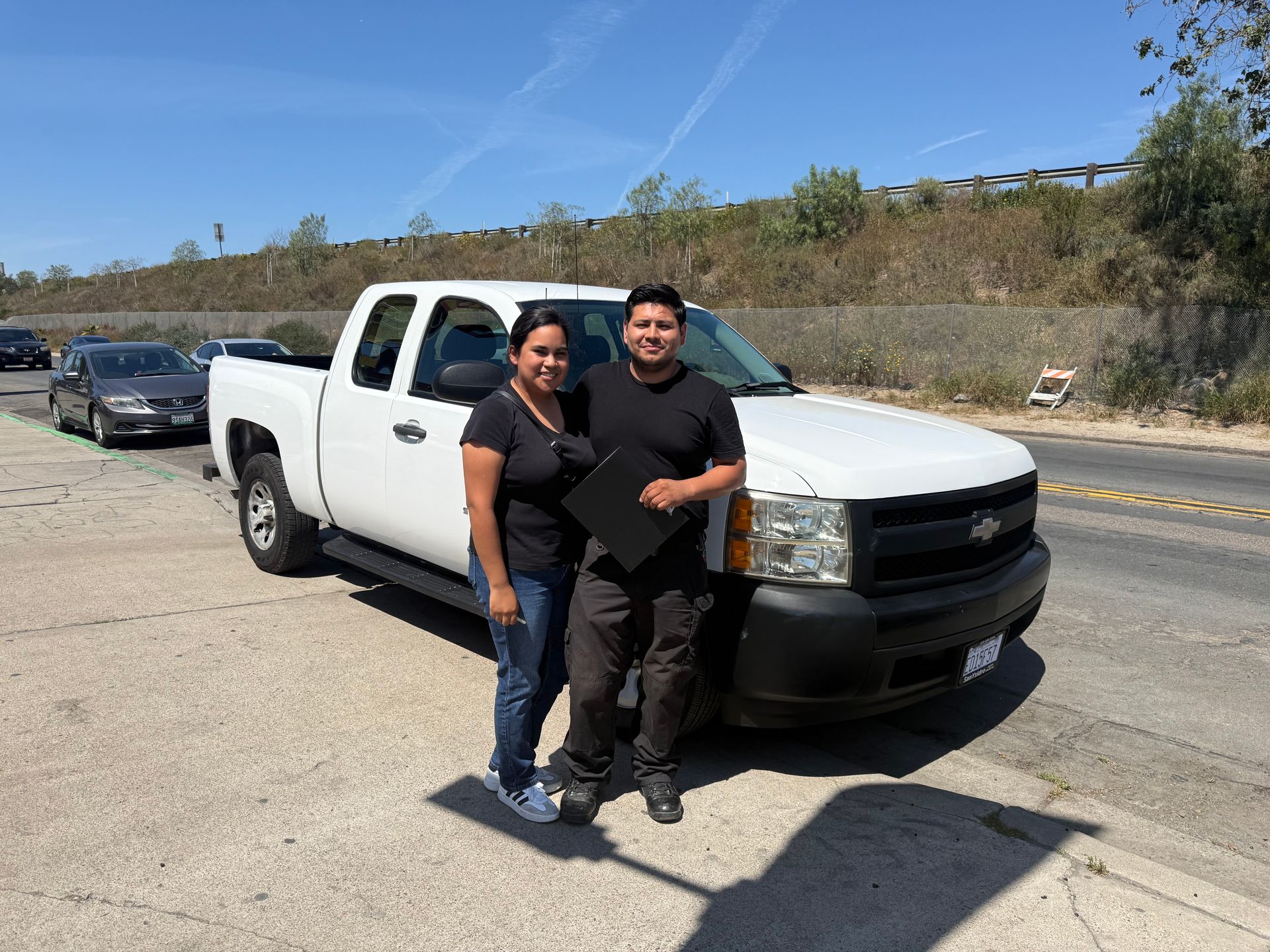 Two people stand in front of a white pickup truck on a roadside under clear skies.