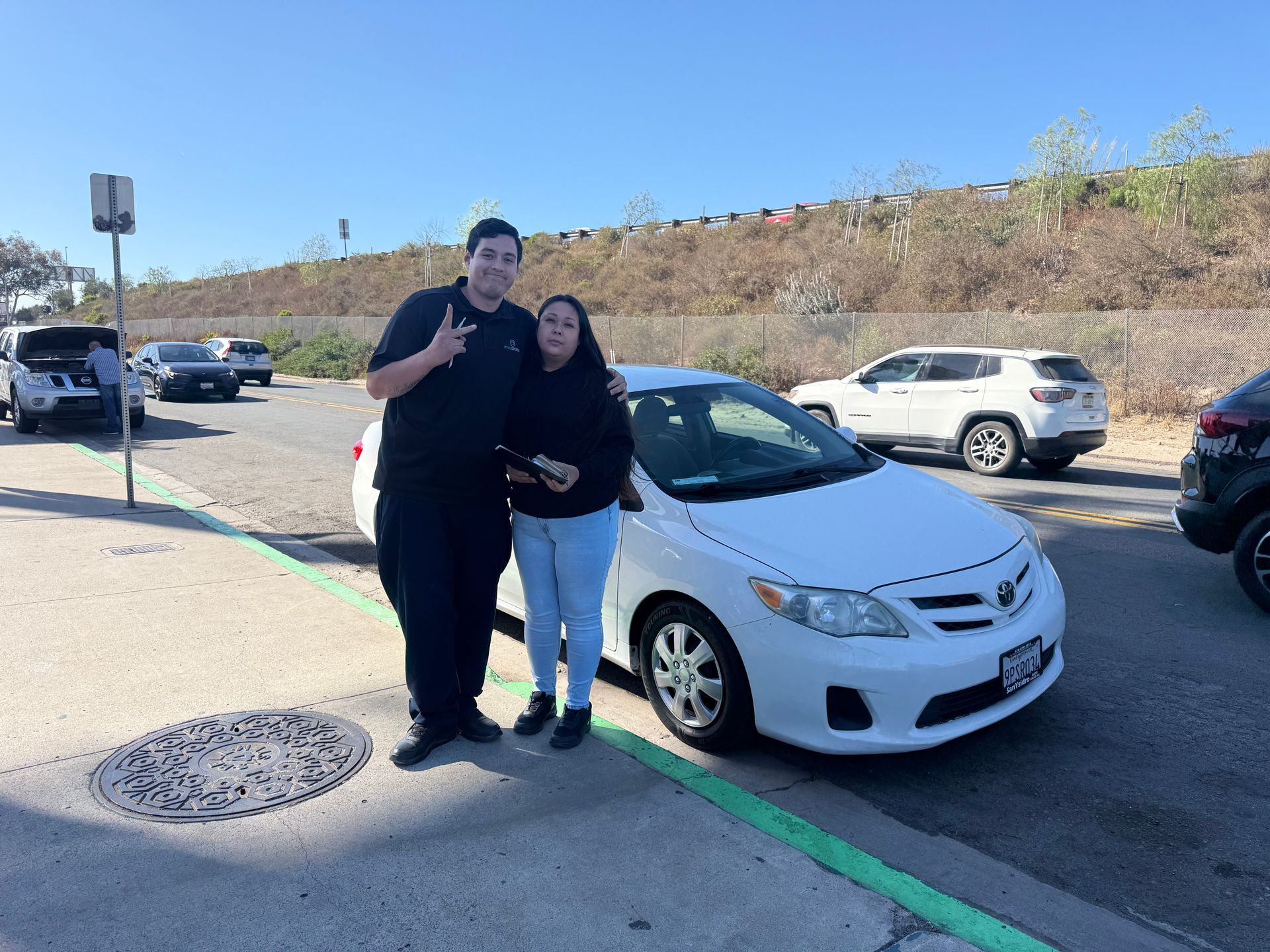 Two people posing beside a white sedan in a sunny parking lot, with other cars and a hillside behind them