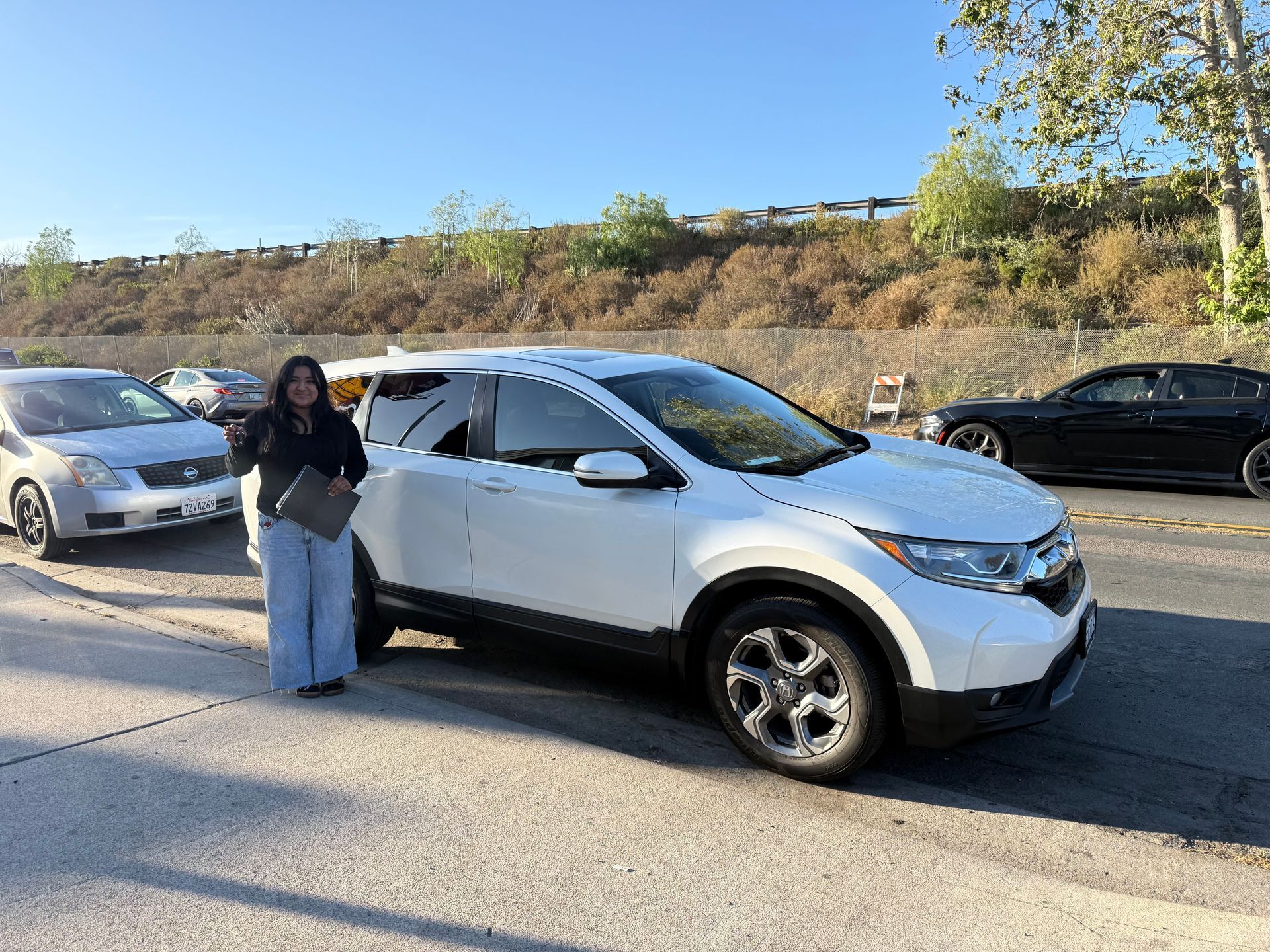 Person standing beside a white SUV on a roadside, with other cars and a hillside in the background.