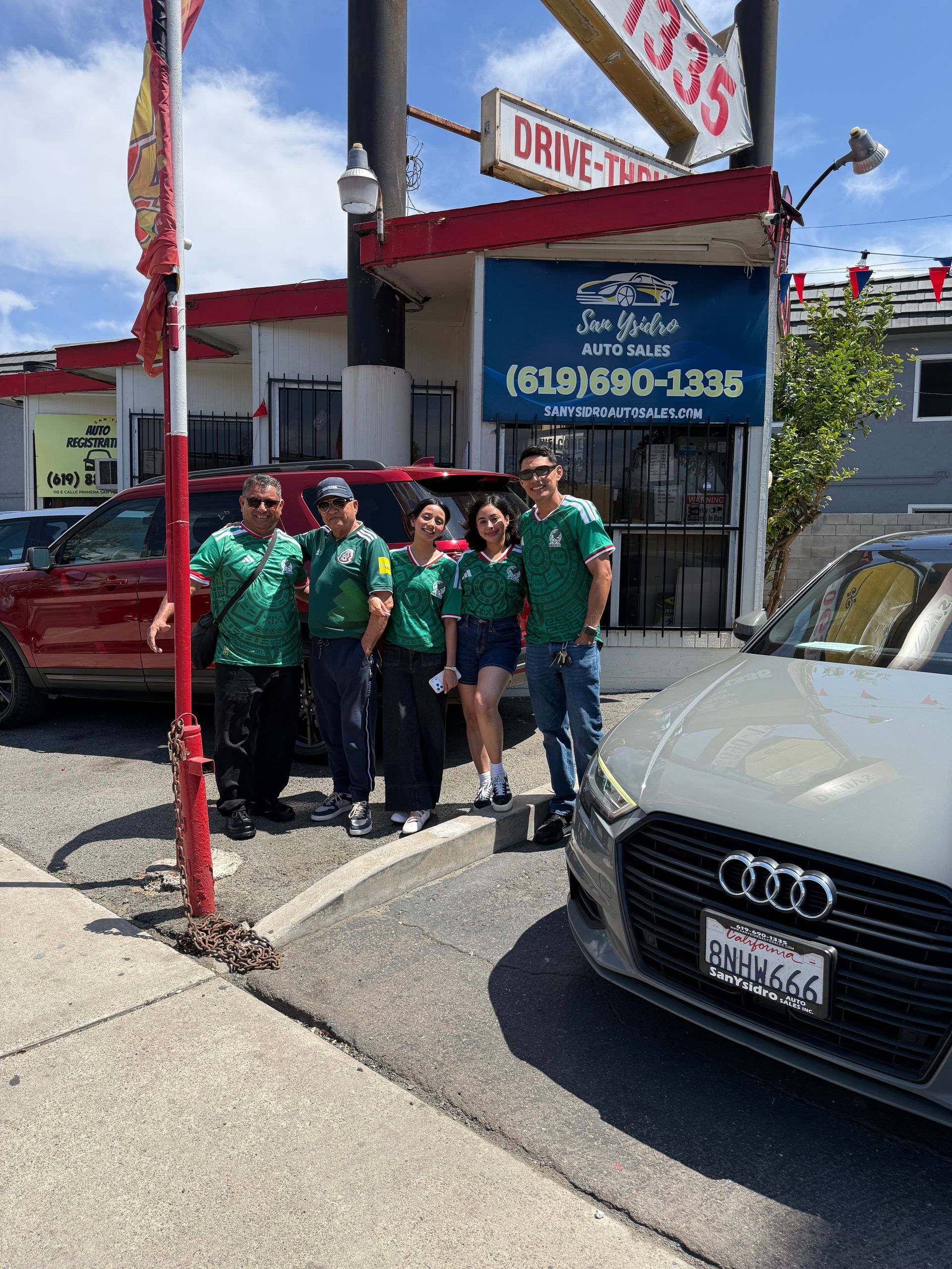 Group of people in green shirts outside a red-and-white storefront beside a parked Audi.