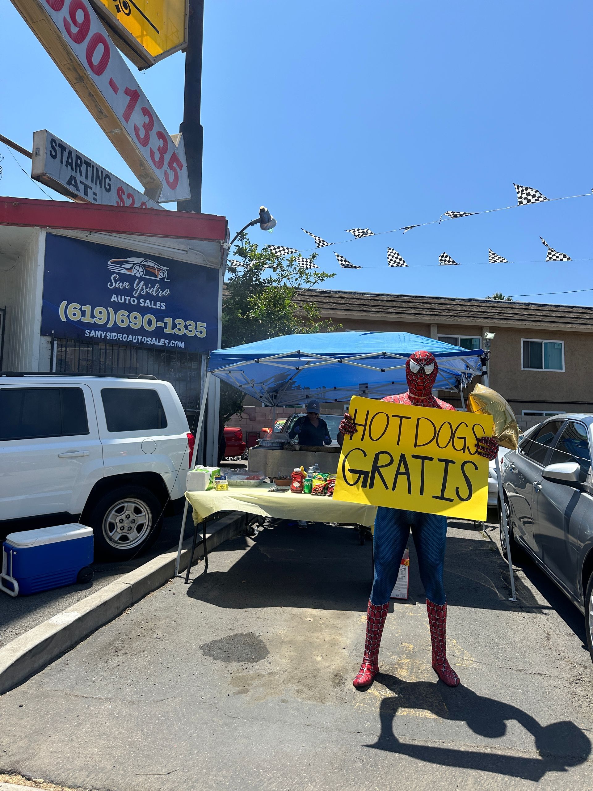 Person holds a yellow sign reading “TUTORING GRATIS” in a sunny parking lot with cars and banners.