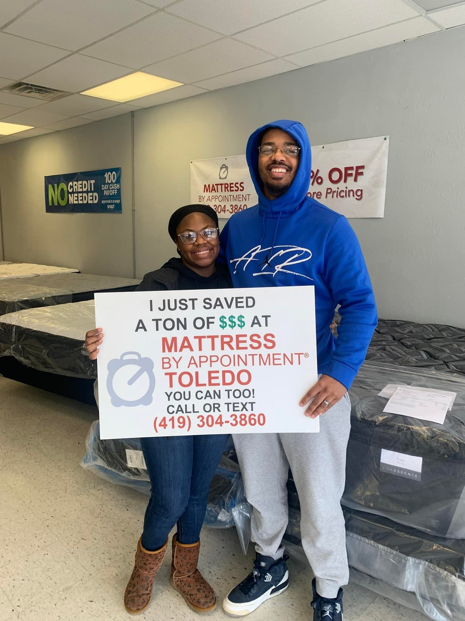 Couple holding a sign, smiling at Mattress By Appointment in Toledo. They stand in a mattress store with mattresses in the background.
