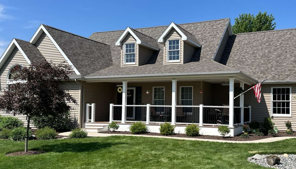 A light-colored house with a porch and dormers, set in a green lawn under a blue sky.