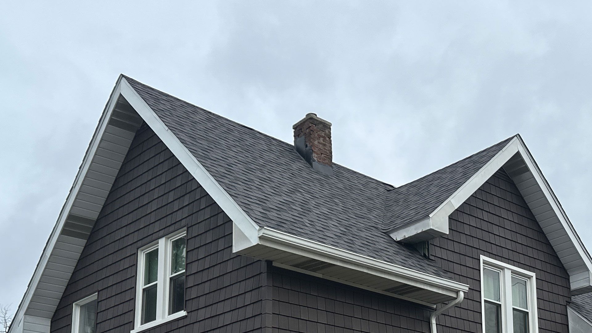 Brown house with gray roof, white trim, and a brick chimney under a cloudy sky.