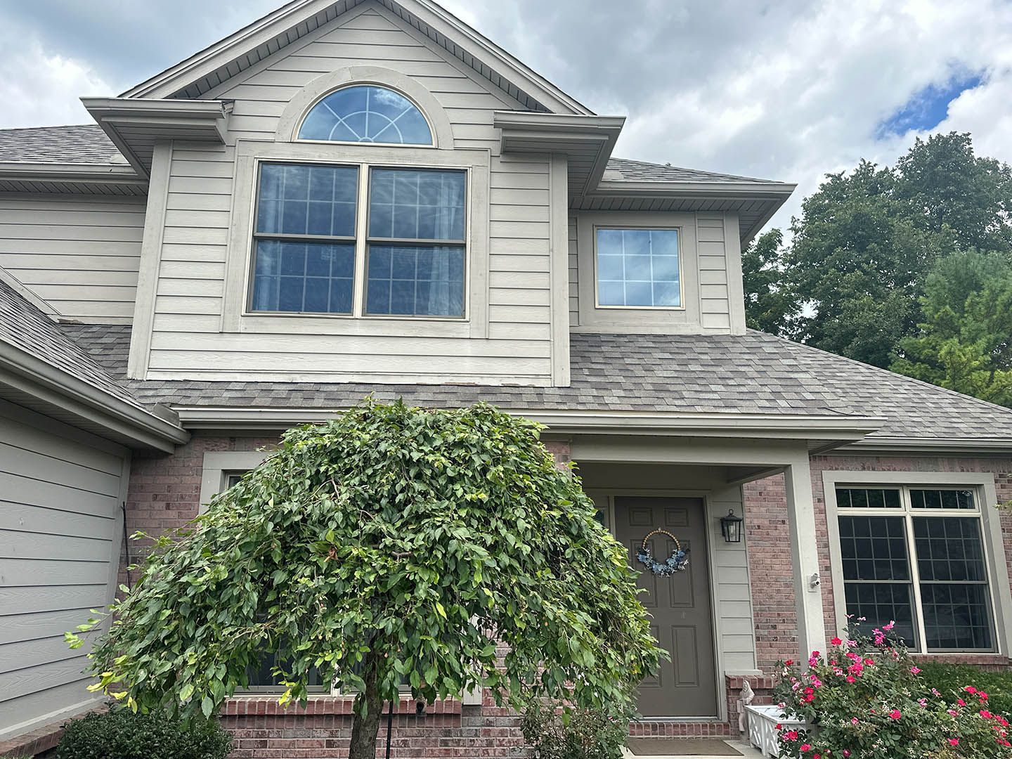 Two-story house with tan siding, brick facade, and a tree in front. Overcast sky.
