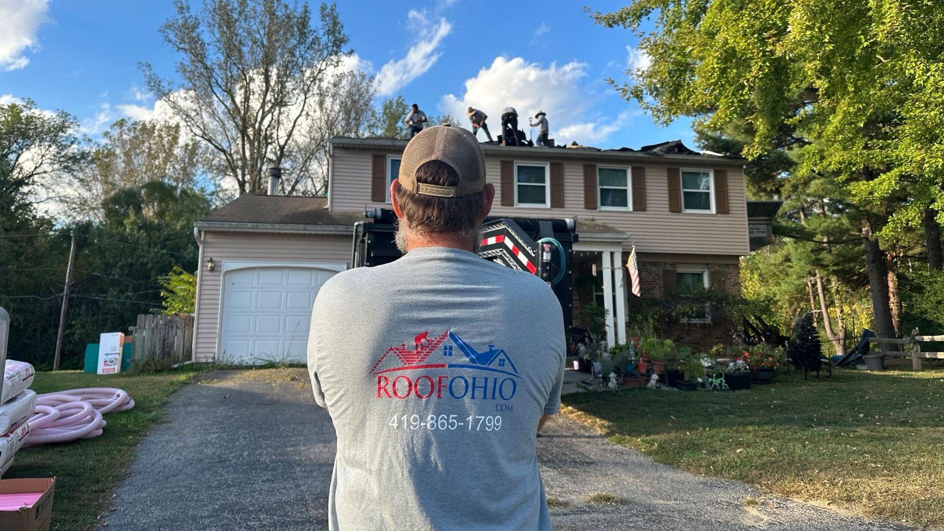 Man facing a house with workers on the roof, likely roofing. Ohio business logo on shirt.