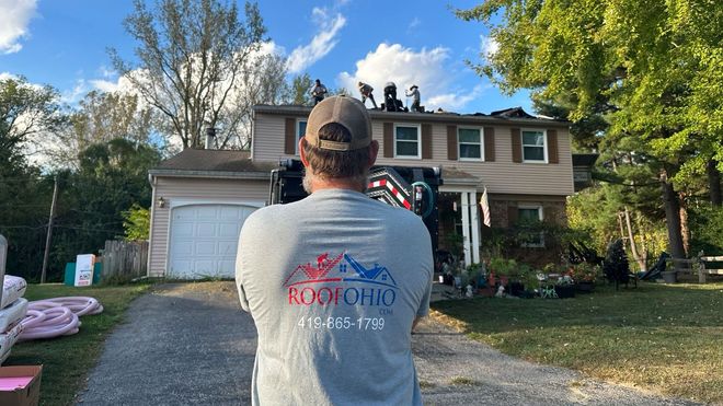 Man facing a house with workers on the roof, likely roofing. Ohio business logo on shirt.