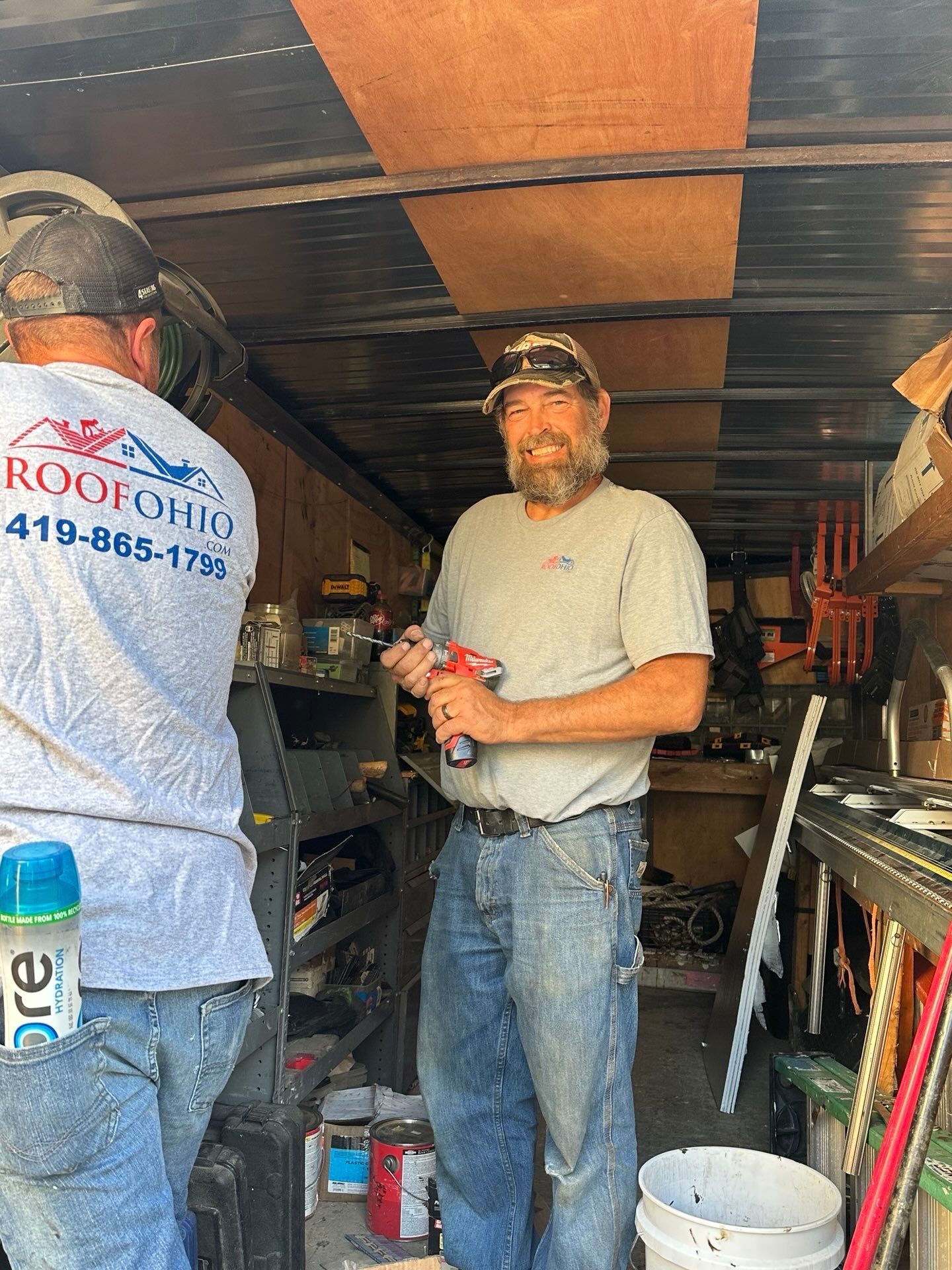 Two men inside a shed with tools. One holds a drill, smiling. The other has a company shirt.