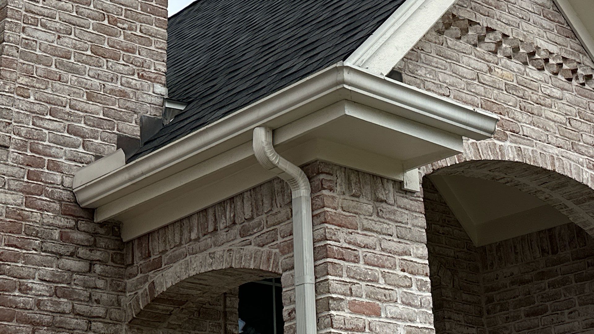 Brick building exterior with beige trim, dark roof, and a gutter system.